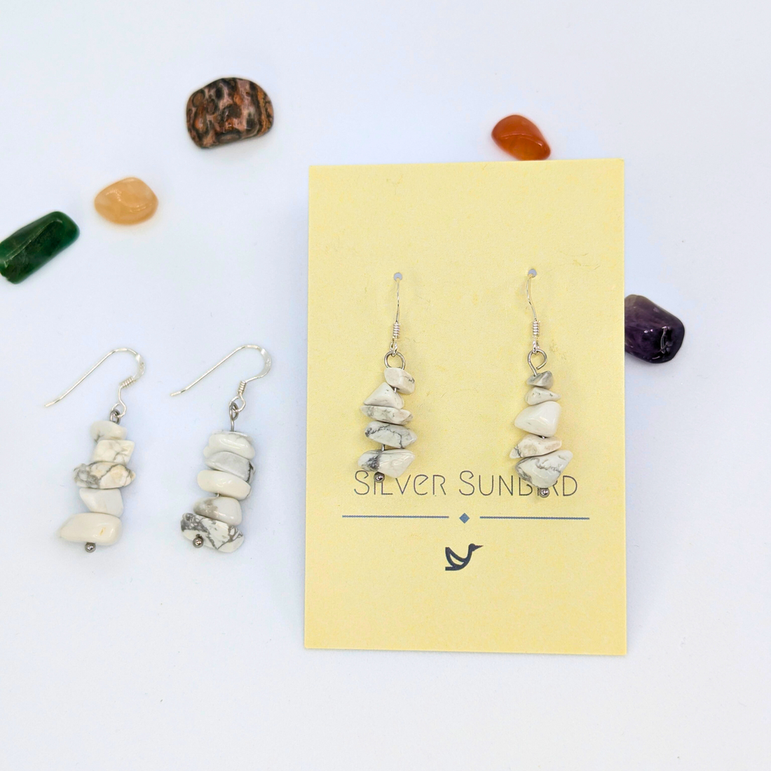 Silver Sunbird White Howlite Gemstone Earrings are displayed on a yellow brand card with another pair to the left. Colorful polished stones surround and highlight these handmade earrings against a white background.