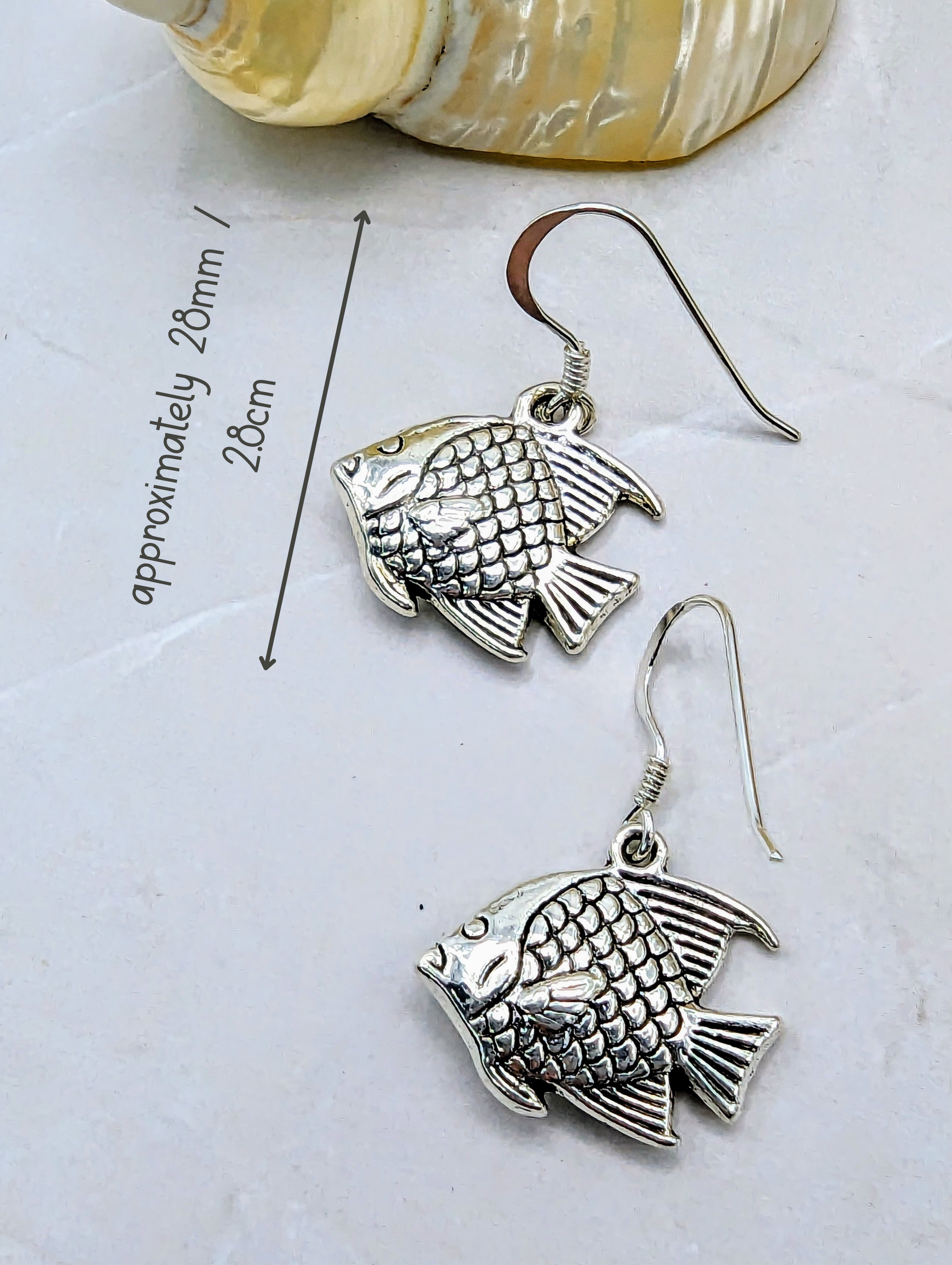 Presenting the Angelic Angelfish Earrings by Silver Sunbird: a pair of sterling silver, fish-shaped dangle earrings, showcasing meticulous scale and fin details. Measuring approximately 28mm in length, these angelfish earrings are artfully displayed next to a measuring line. A seashell can be seen partially in the background on a light-coloured surface—an ideal choice for any ocean enthusiast.
