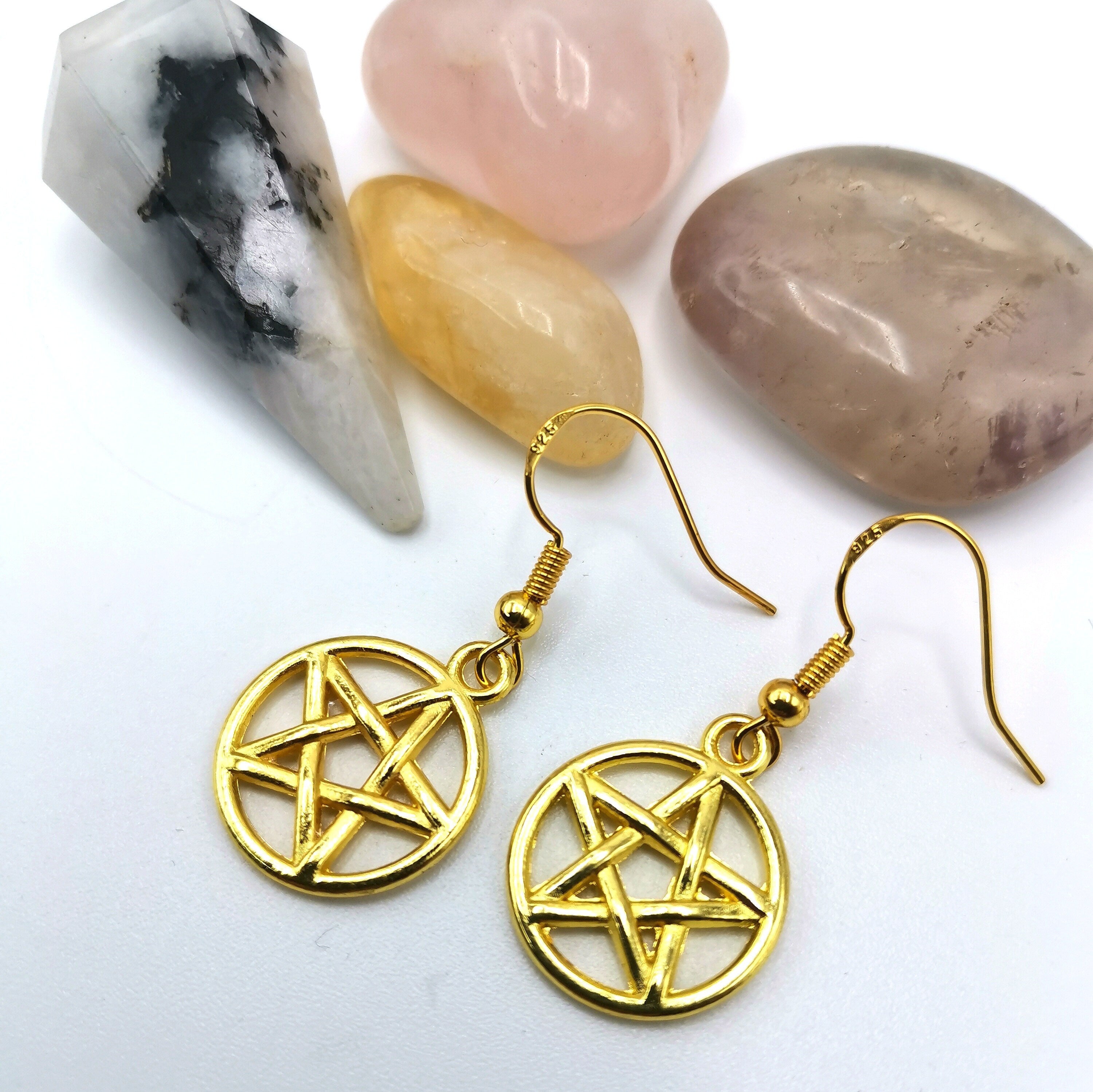 Silver Sunbird’s Pentagram Earrings are shown on a white surface, with rose quartz, amethyst, yellow citrine, and a white-black speckled stone arranged behind them to highlight their pagan-inspired design.