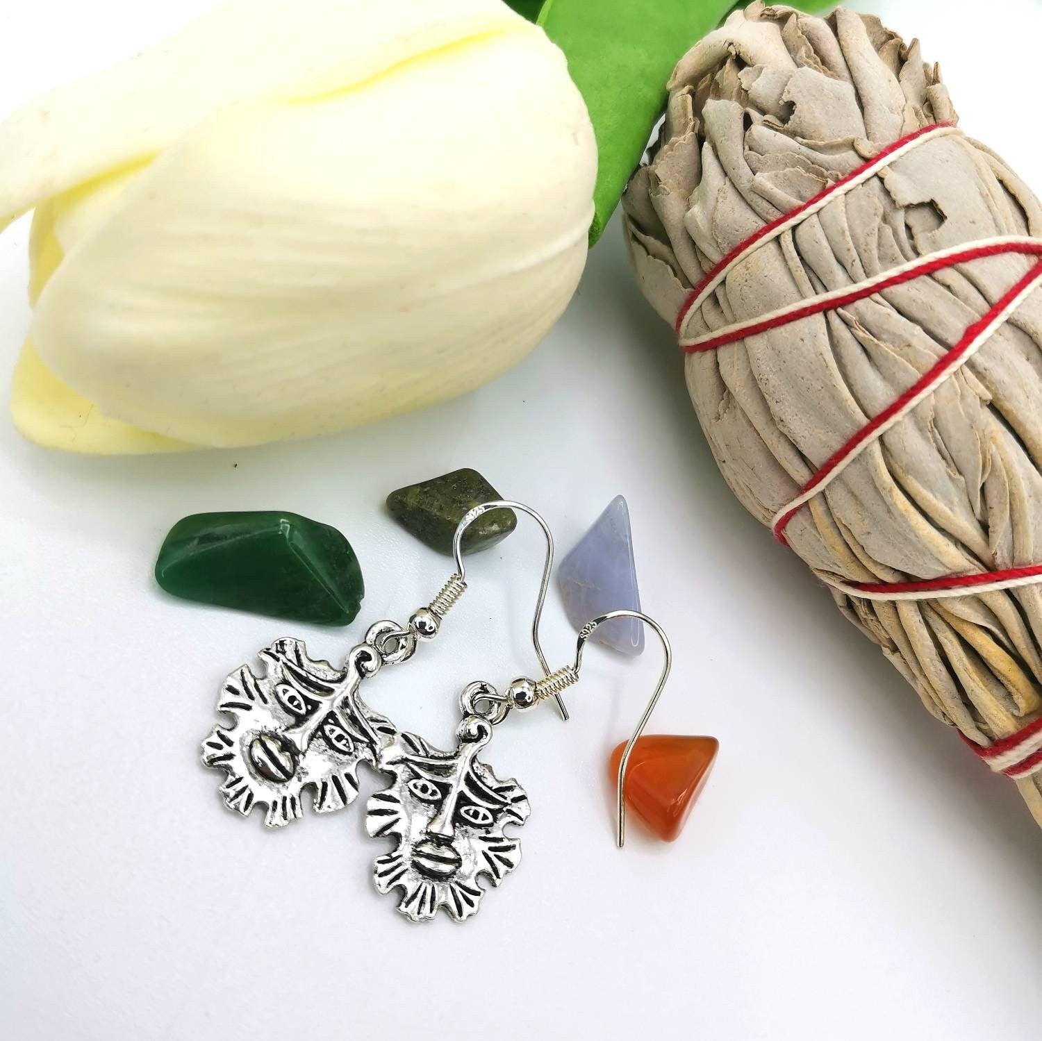 The Gentle Green Man Silver Earrings by Silver Sunbird, crafted from 925 sterling silver with intricate pagan designs, are nestled among colorful stones, a large white flower, and a sage bundle tied with red string on a pristine white surface.