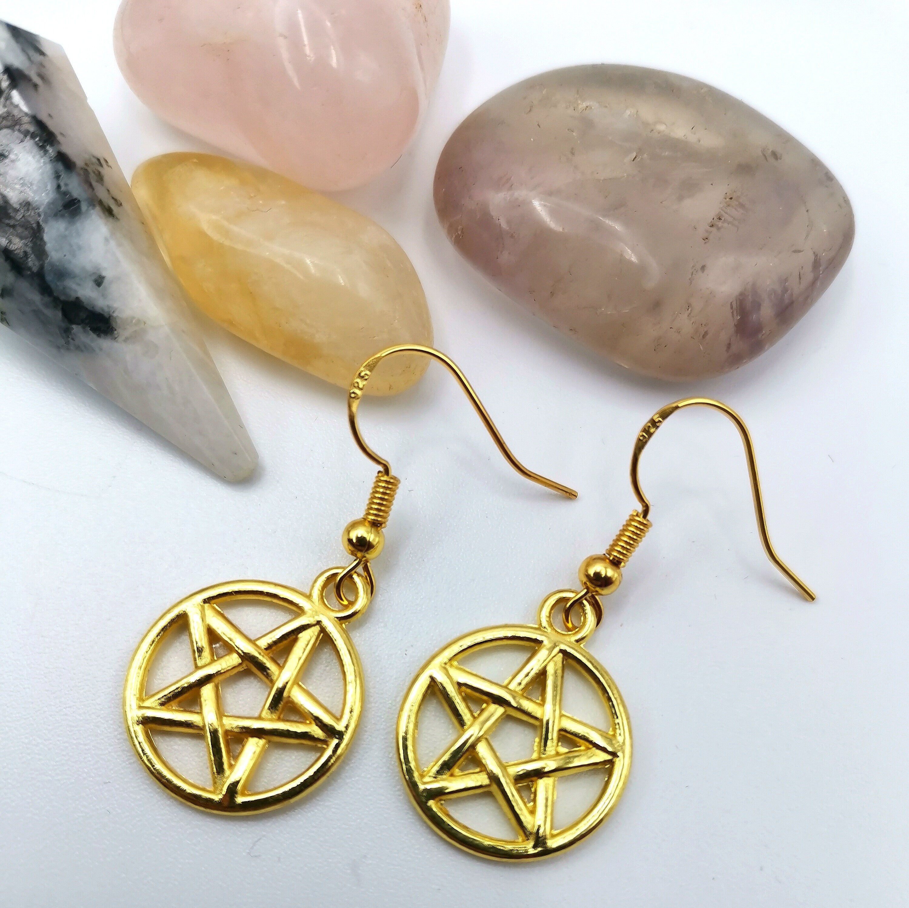 Pentagram Earrings by Silver Sunbird, symbolizing pagan heritage, are displayed on a white surface next to four polished crystals in pink, yellow, gray, and white with black streaks.