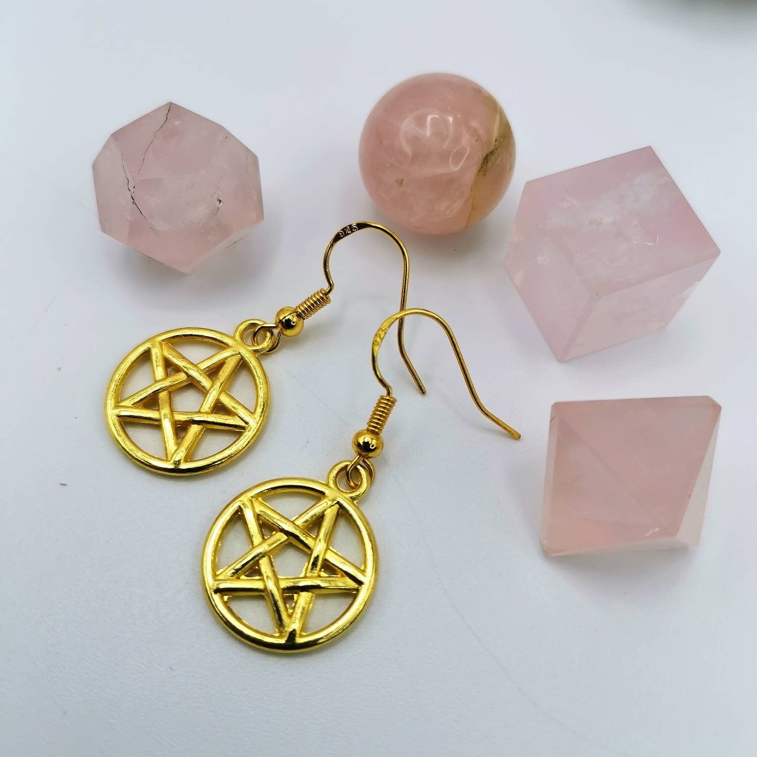 Silver Sunbird Pentagram Earrings are displayed on a white surface, surrounded by four rose quartz stones in geometric shapes—a sphere, cube, and two prisms.