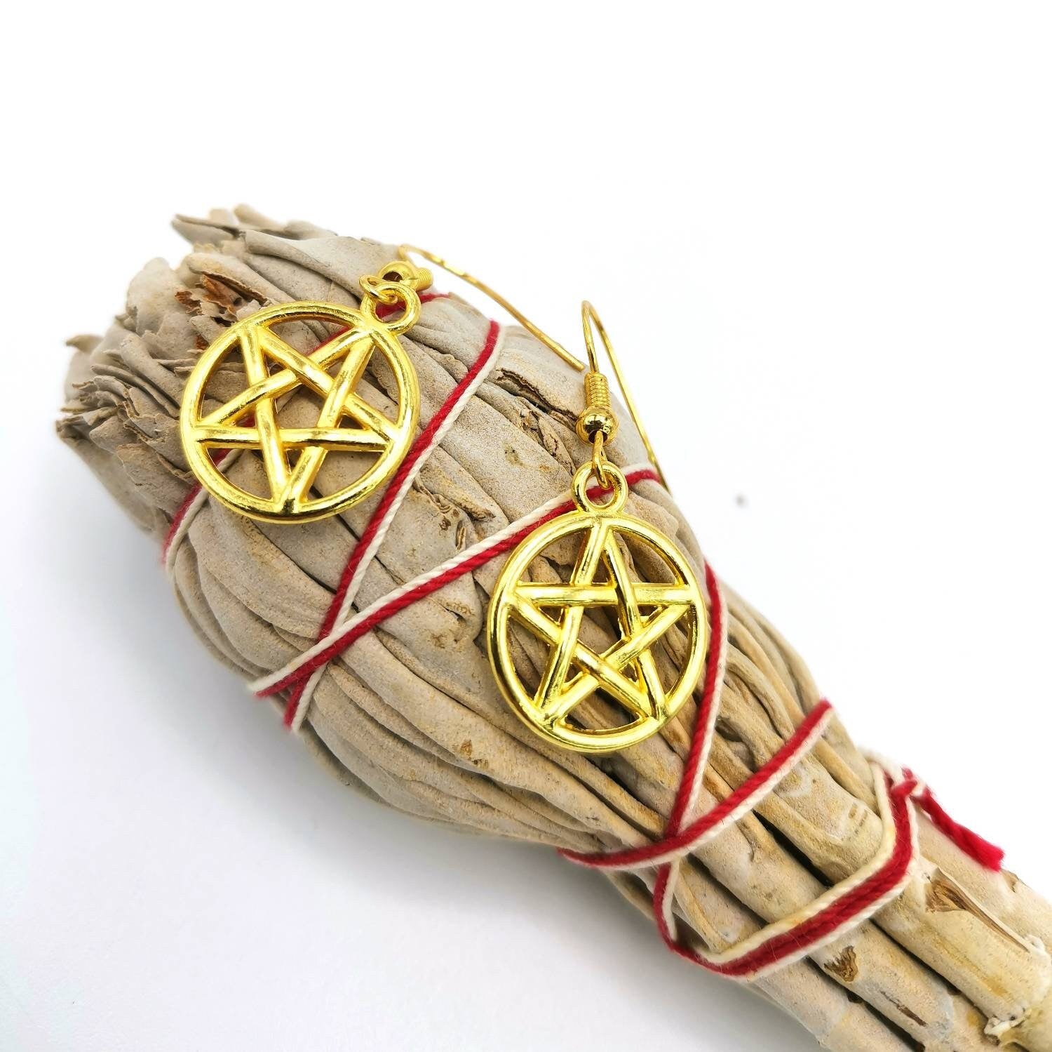 A bundle of dried sage tied with red string is topped with Silver Sunbird Pentagram Earrings, set against a white background.