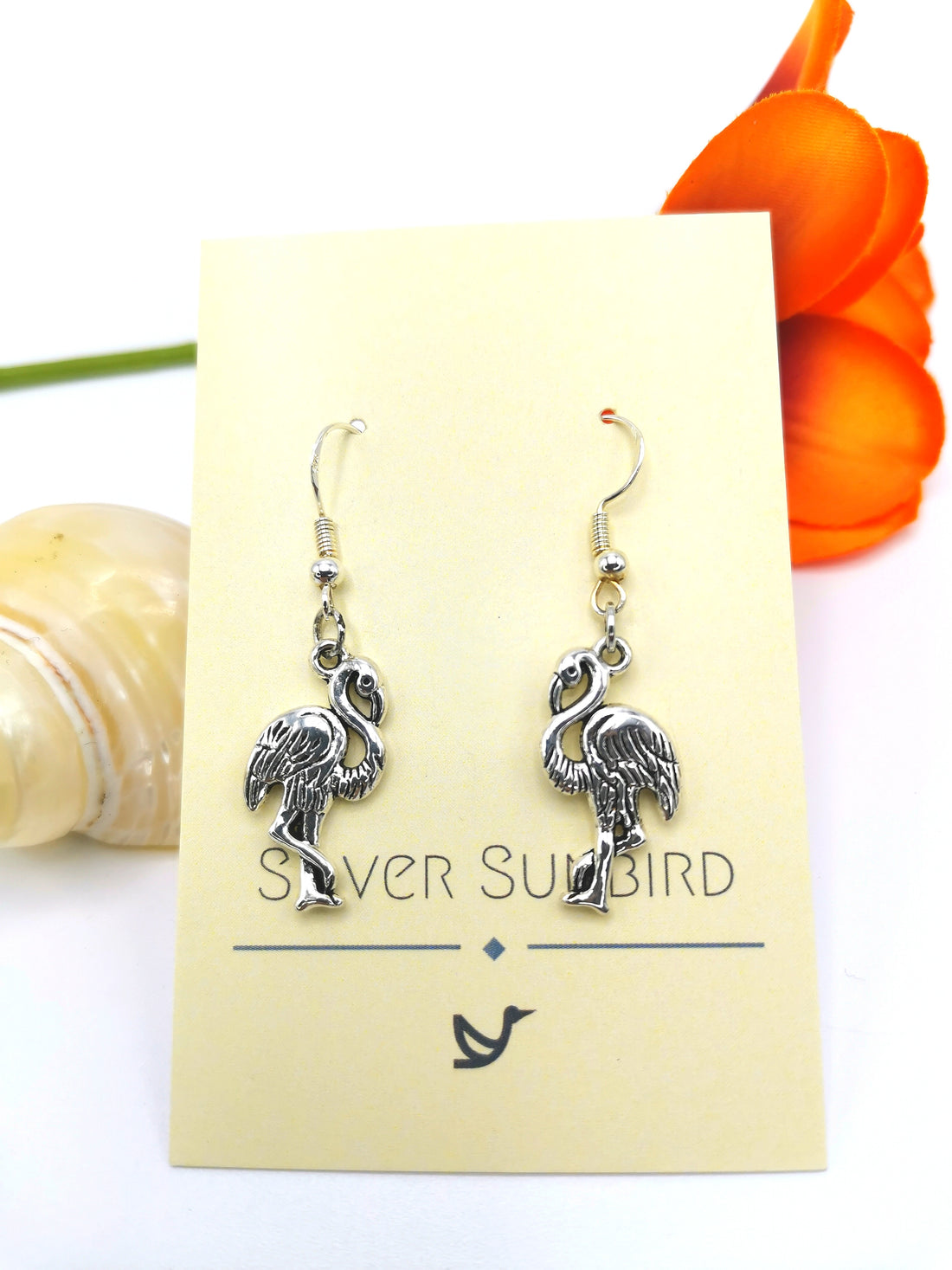 A pair of Flocking Flamingo Earrings is displayed on a beige card with "Silver Sunbird" printed on it. An orange flower and a seashell are placed beside the card. The handmade jewelry features intricate detailing of standing birds, crafted from 925 sterling silver.