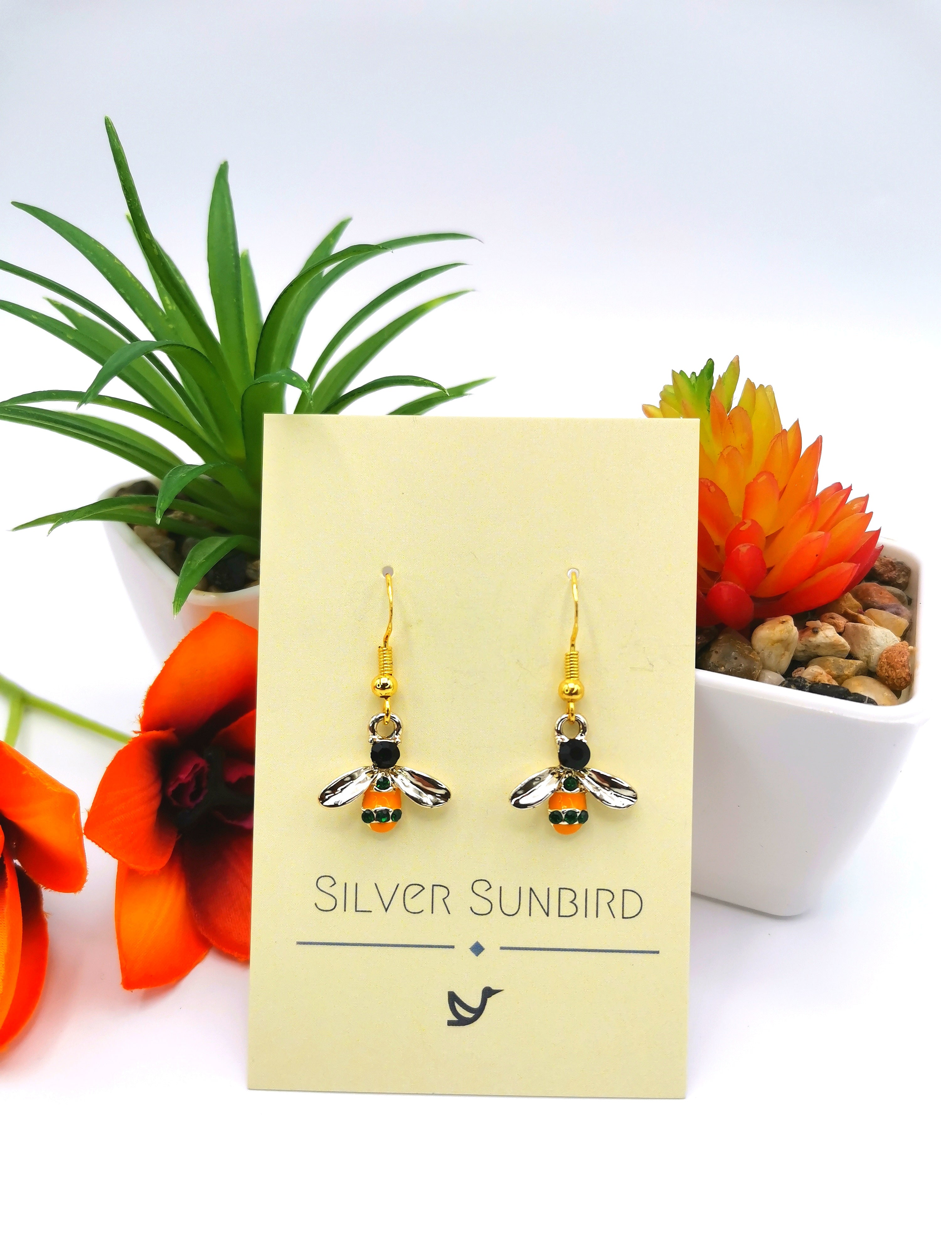 The Bee Happy Bee Earrings, an exquisite pair of handmade decorative drop earrings featuring black, silver, and orange stones, are beautifully presented on a Silver Sunbird card. These earrings are accented by sterling silver and perfectly complemented by vibrant orange flowers and a succulent in white pots.