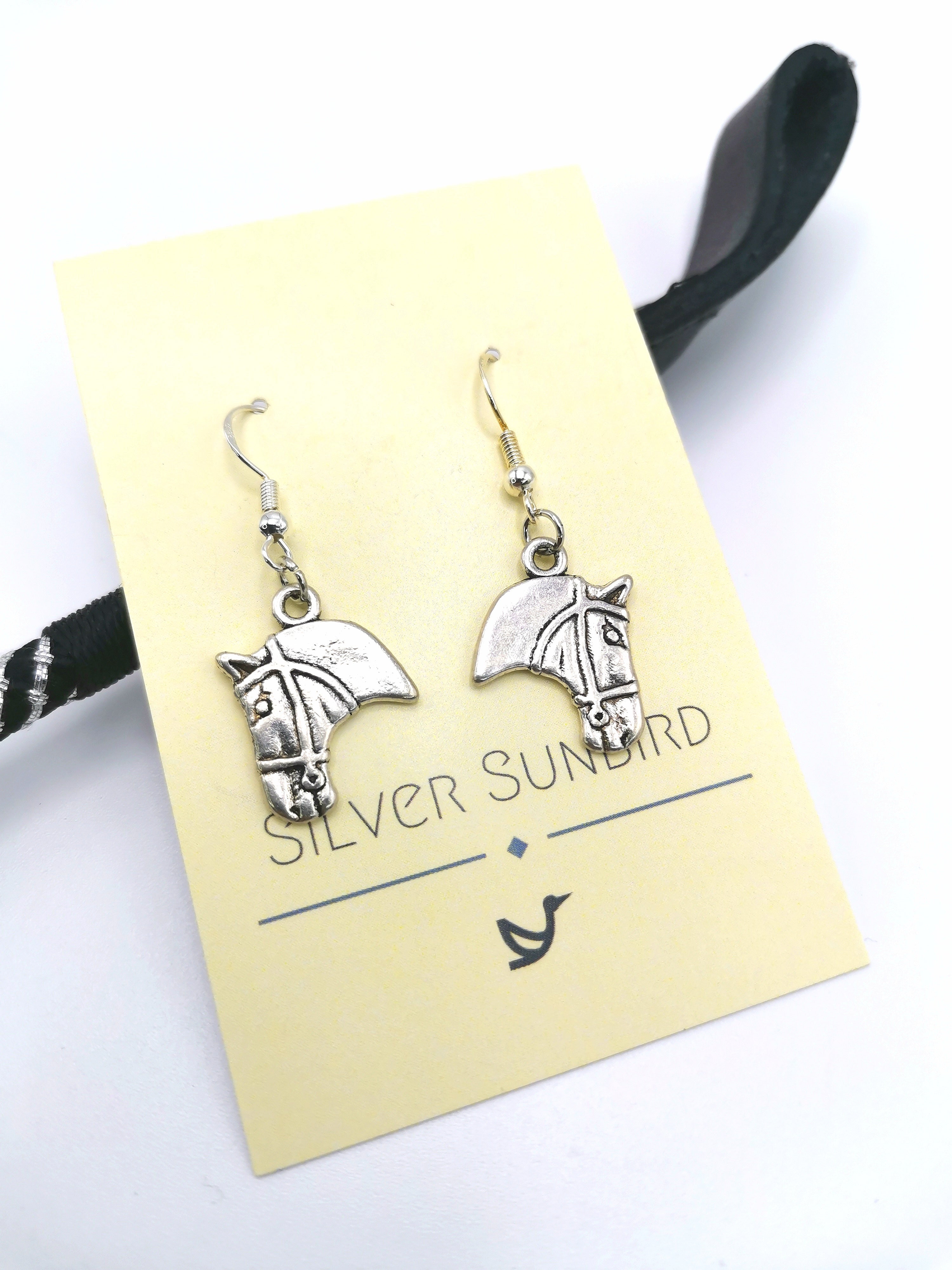 The Take The Reins Equine Earrings, crafted from sterling silver and ideal for any horse lover, are beautifully presented on a yellow card labeled "Silver Sunbird." A black ribbon can be partially seen adjacent to the card.