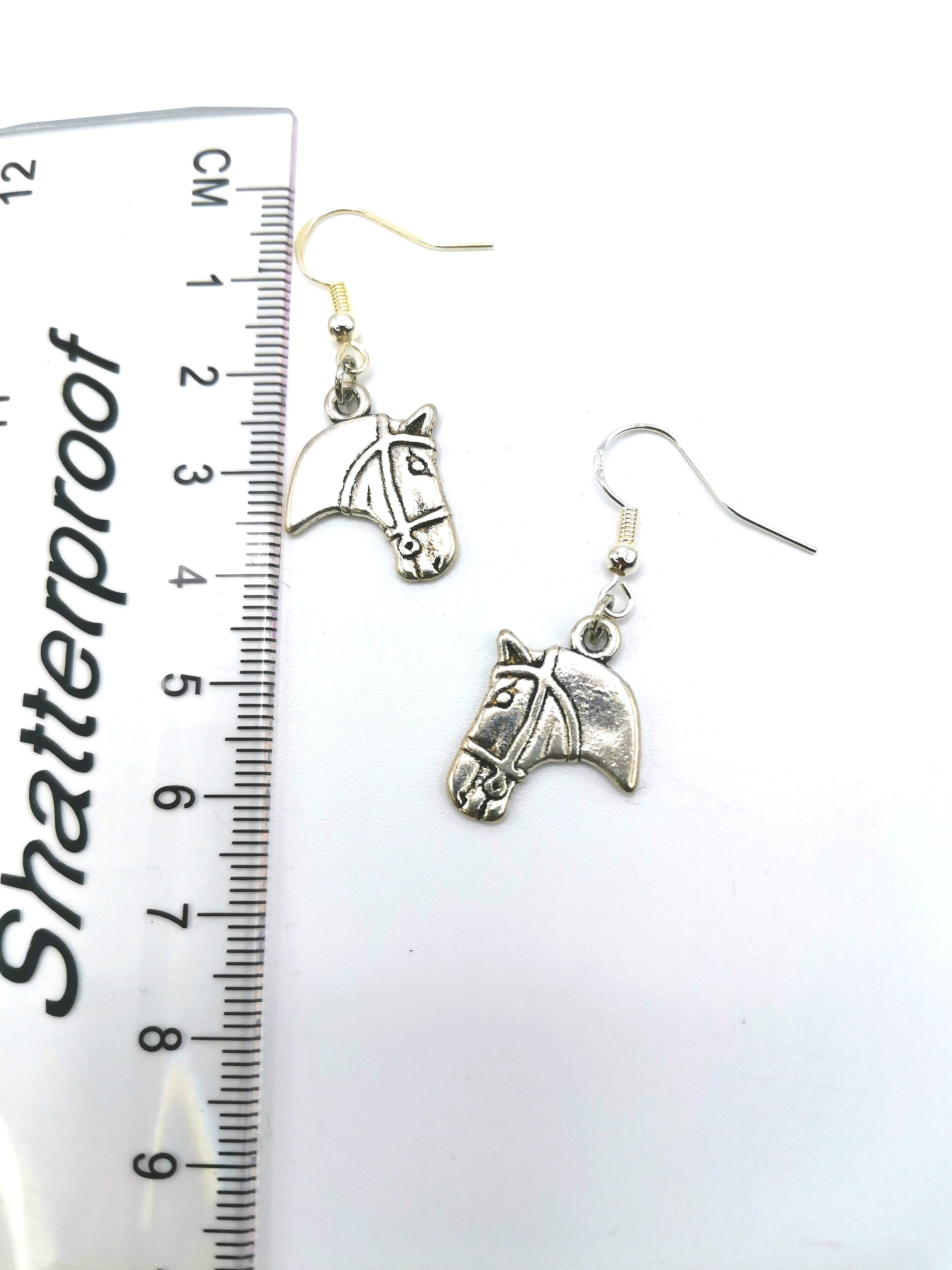 The Take The Reins Equine Earrings by Silver Sunbird, featuring sterling silver horse heads with hooks, are displayed alongside a ruler marked in centimeters and inches to highlight the equestrian charm of this handcrafted jewelry.