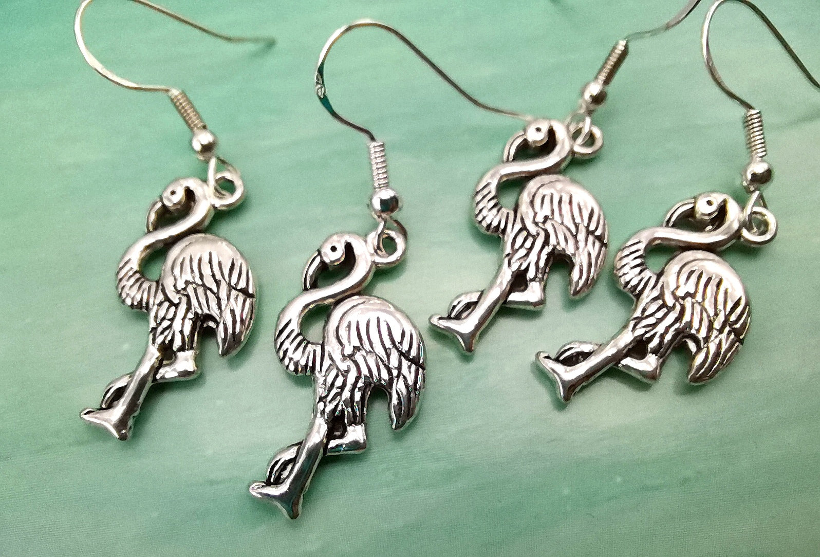 Two "Flocking Flamingo Earrings" from Silver Sunbird are displayed against a greenish background. Each piece of this handmade jewellery showcases a detailed flamingo design with one leg extended forward, highlighting the intricate feather details and curved neck. Crafted from 925 sterling silver, they exude elegance.