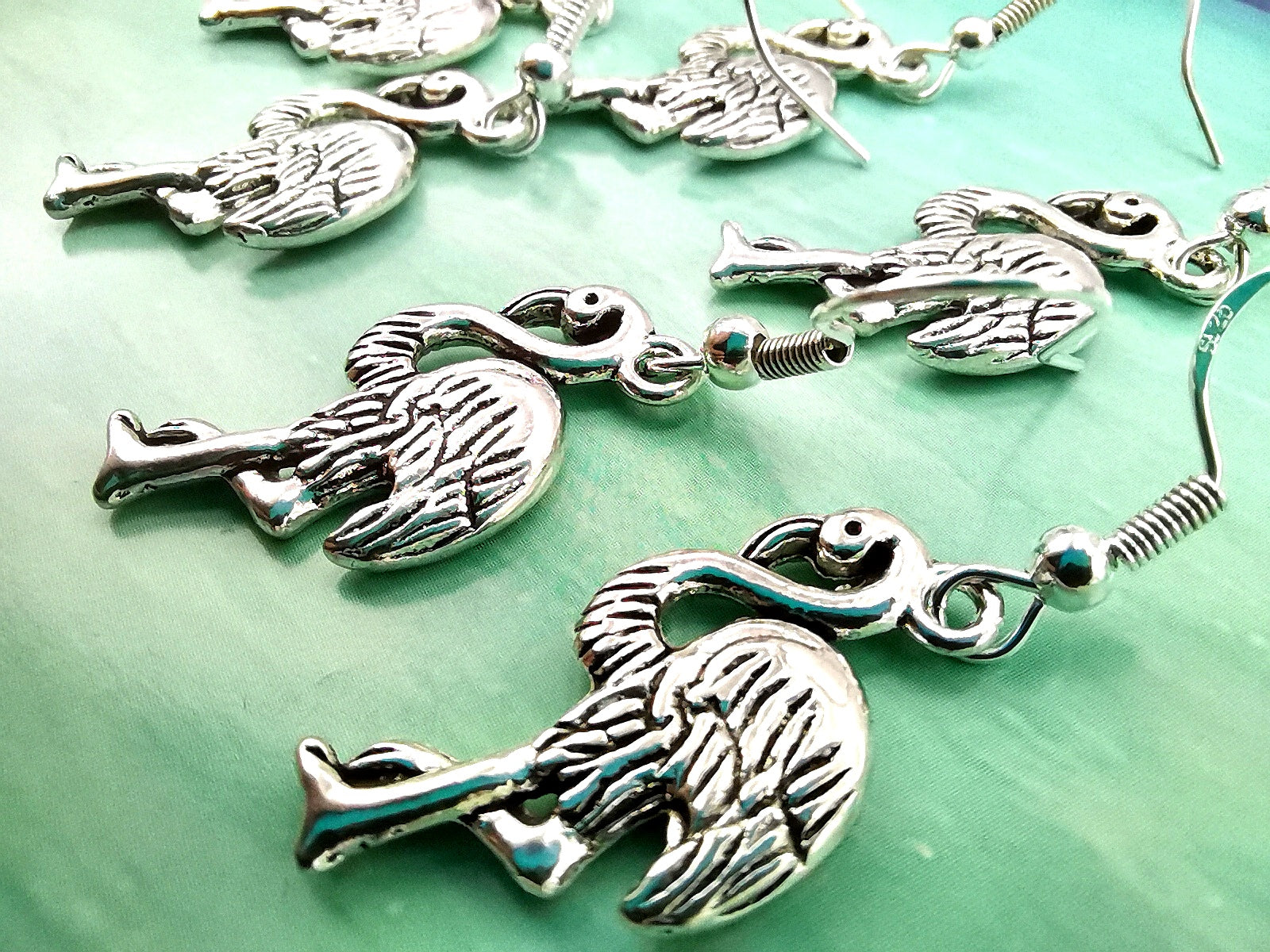 A close-up of multiple pairs of Flocking Flamingo Earrings from Silver Sunbird, arranged on a green and blue gradient background. These 925 sterling silver earrings feature intricate feather details and are connected to small hooks for wearing, showcasing the beauty of handmade jewellery.