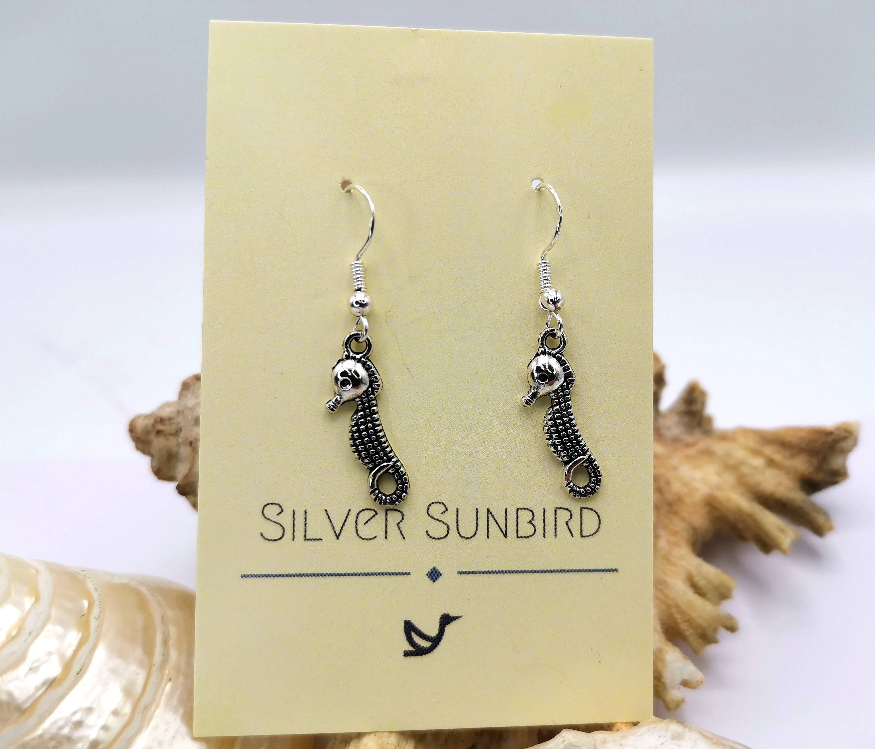 The Spirited Seahorse Earrings by Silver Sunbird, crafted from 925 sterling silver, are elegantly displayed on a branded card amidst a backdrop of seashells.