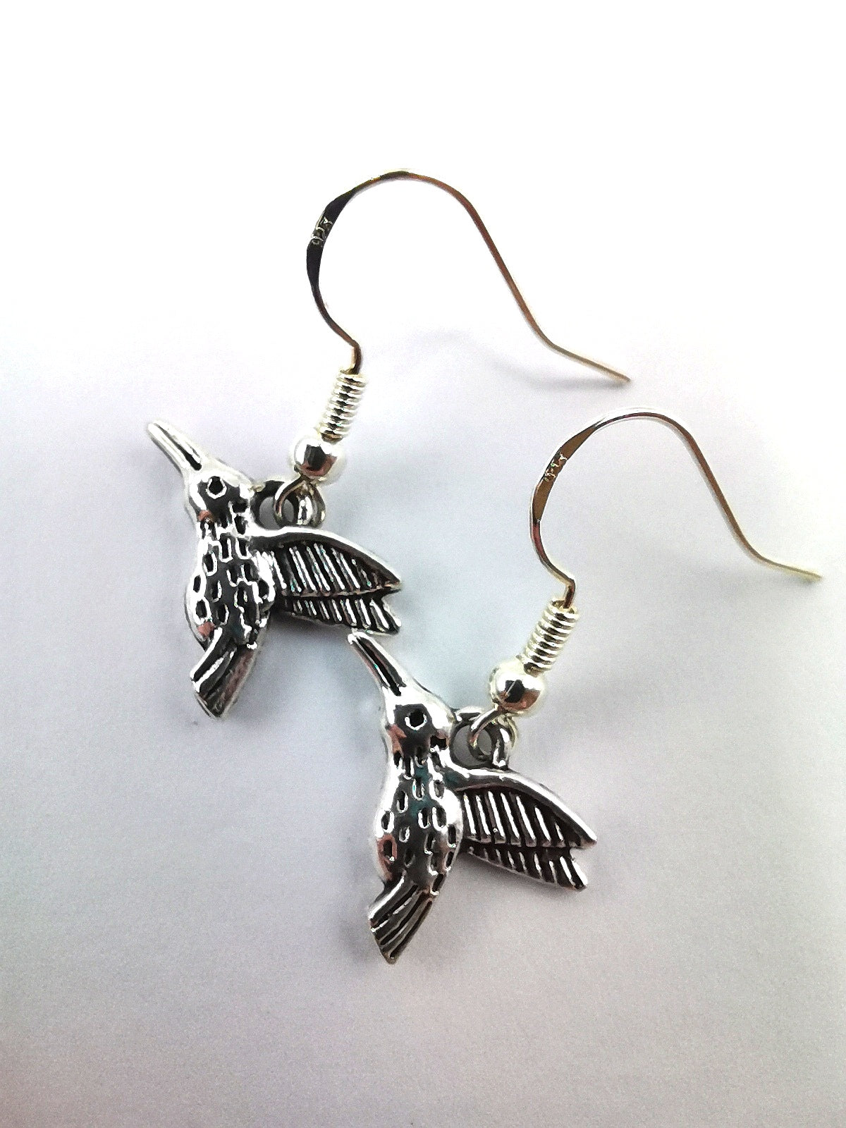 A pair of Heartfelt Hummingbird Earrings by Silver Sunbird, made from sterling silver with intricate detailing and fishhook backings. The birds&