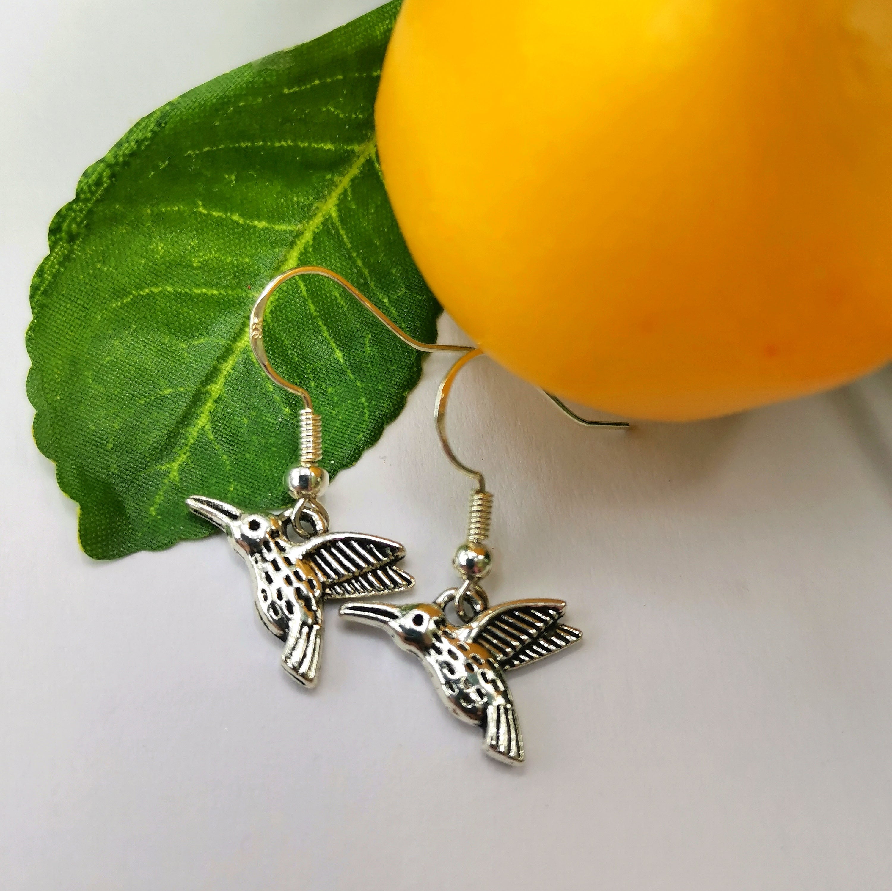 A pair of Heartfelt Hummingbird Earrings by Silver Sunbird are placed against a backdrop of a green leaf and a yellow fruit. The detailed handmade jewelry captures the birds in mid-flight with outspread wings. The green leaf and yellow fruit add a natural, vibrant touch to the image.