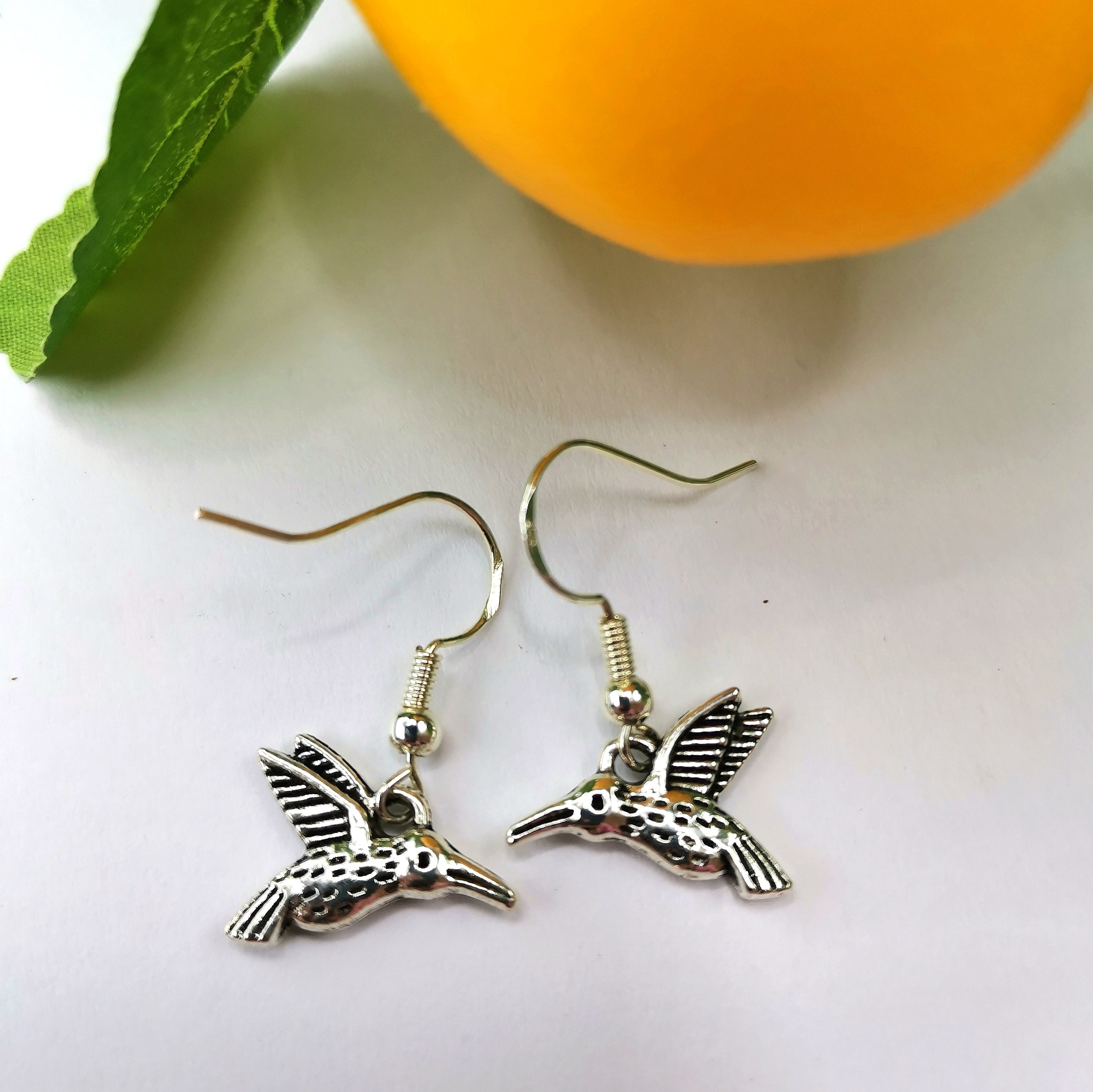 The Silver Sunbird Heartfelt Hummingbird Earrings, a pair of handmade, sterling silver earrings with hook fastenings, are displayed on a white surface. Positioned next to an orange fruit with a green leaf attached, the earrings create a bright and cheerful composition.