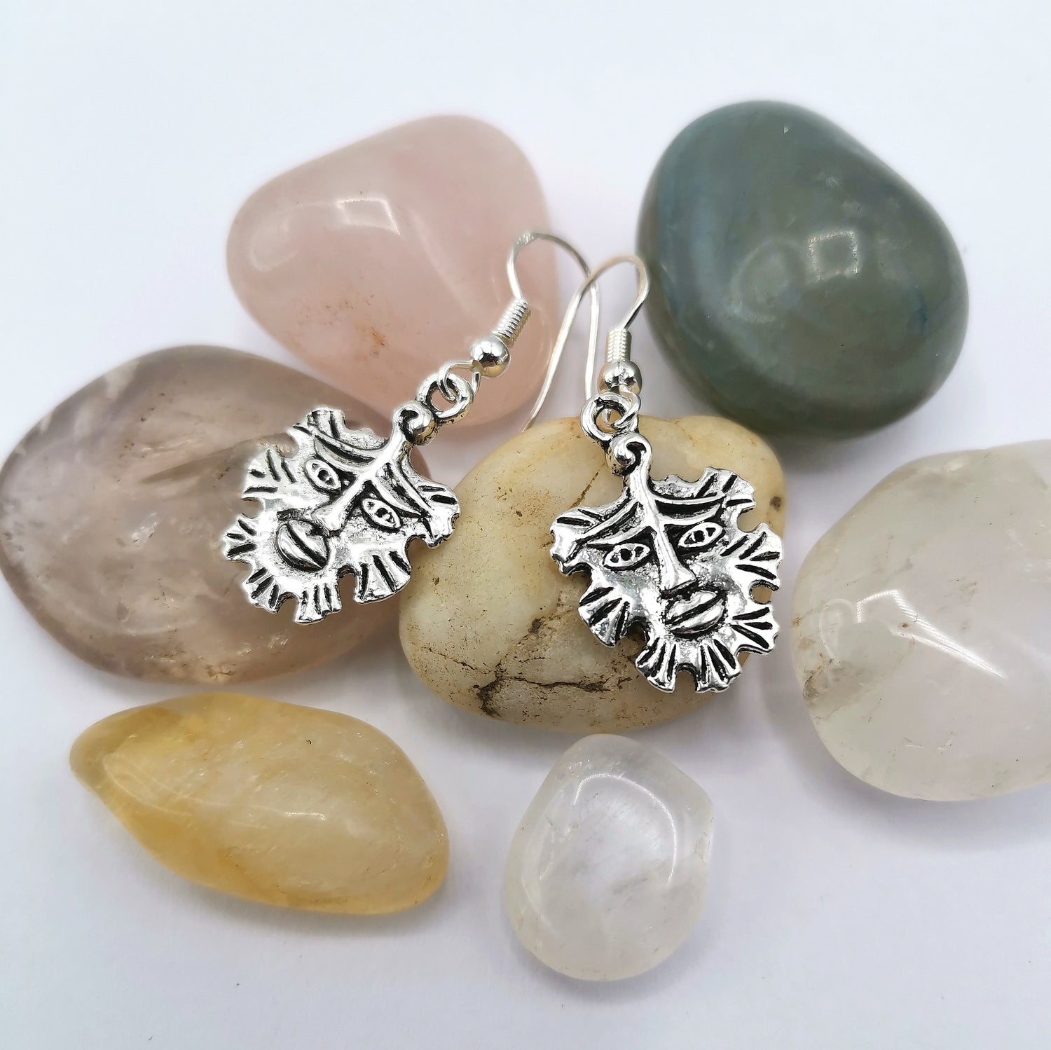 The Gentle Green Man Sterling Silver Earrings by Silver Sunbird feature sun-like faces and rest on smooth stones in pink, green, yellow, and clear tones against a white backdrop—a striking piece of pagan jewelry.