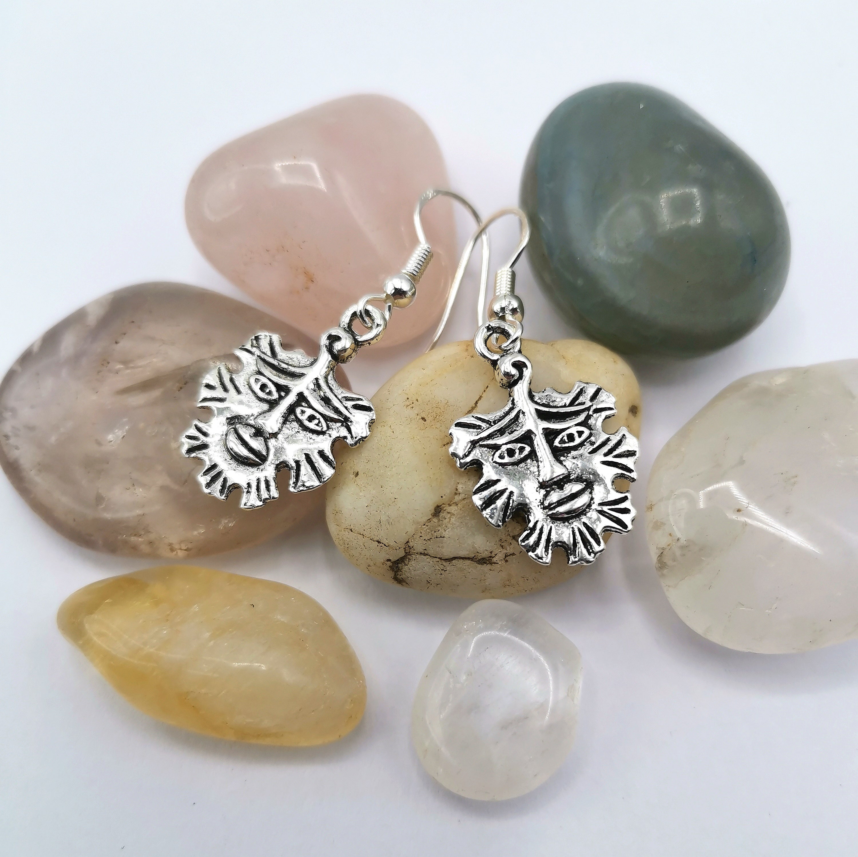 The Gentle Green Man Sterling Silver Earrings by Silver Sunbird feature sun-like faces and rest on smooth stones in pink, green, yellow, and clear tones against a white backdrop—a striking piece of pagan jewelry.