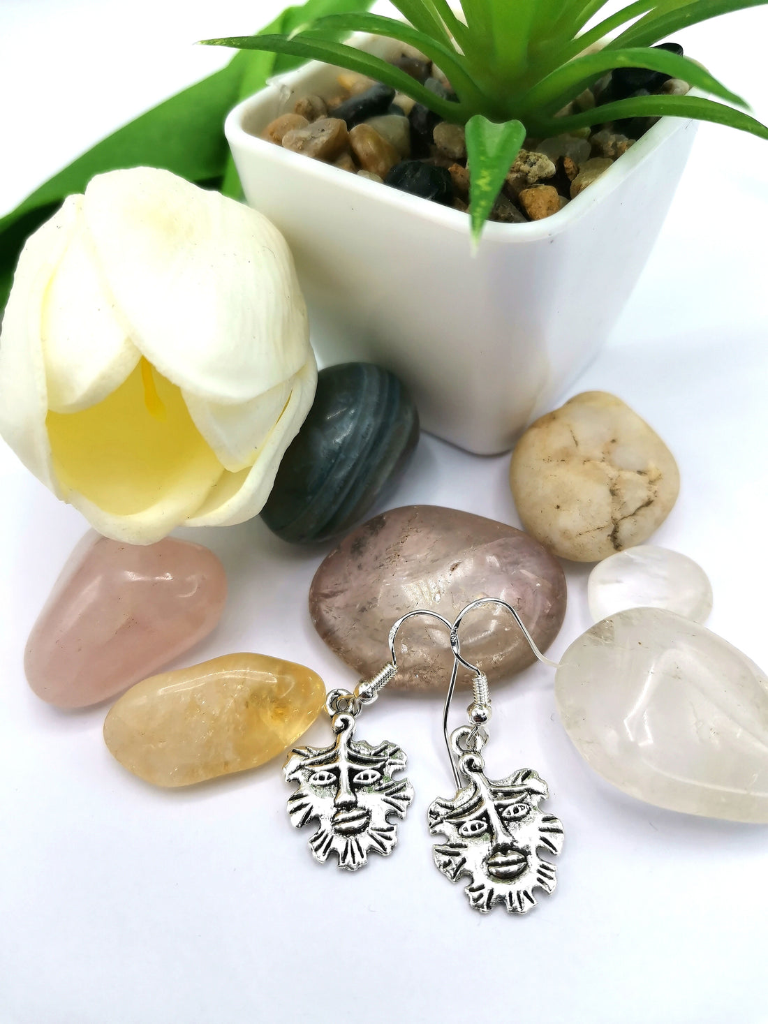 The Gentle Green Man Sterling Silver Earrings by Silver Sunbird are displayed on polished stones beside a white tulip and a small potted succulent, making them perfect for fans of unique pagan jewellery.