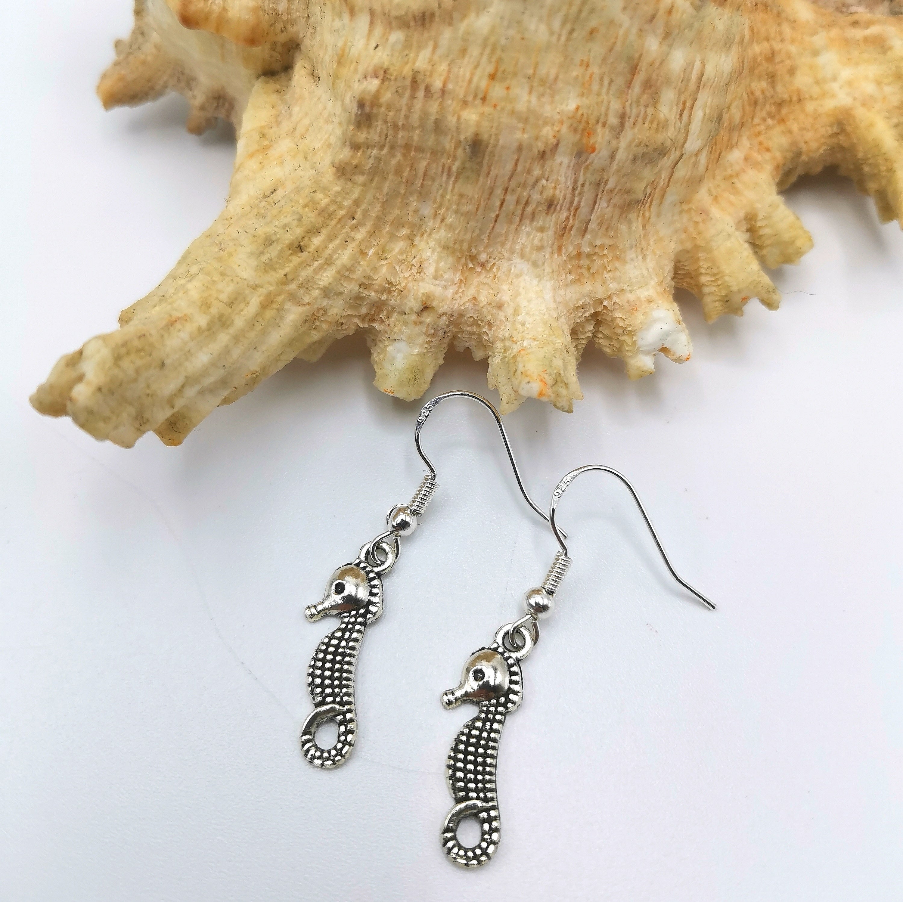 The Spirited Seahorse Earrings by Silver Sunbird, made from 925 sterling silver, are elegantly showcased against a large, textured seashell on a white background. The intricate detailing on the earrings perfectly complements the natural patterns of the shell.