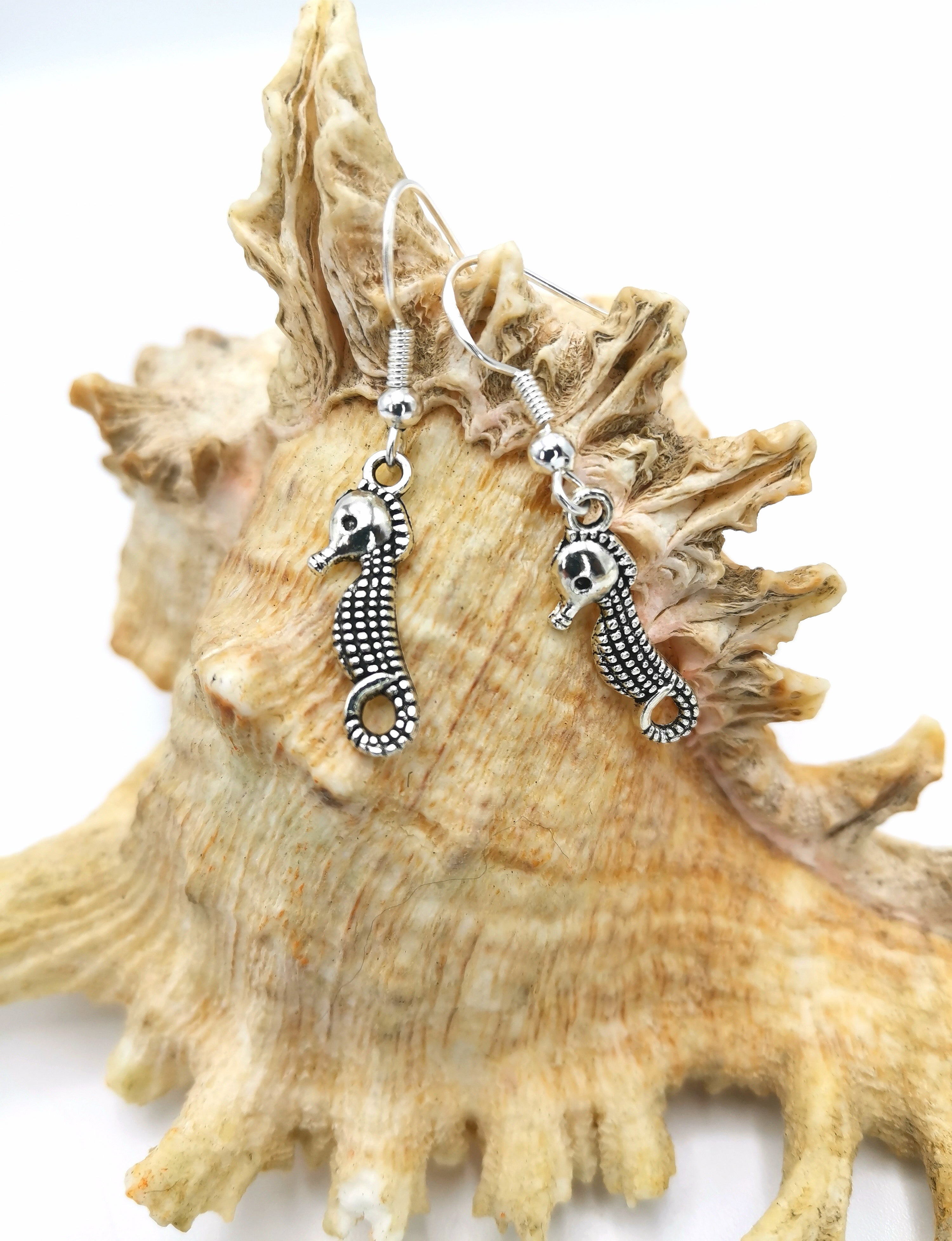 Spirited Seahorse Earrings from Silver Sunbird are showcased on a textured seashell. The detailed design of the 925 sterling silver seahorses stands out against the natural surface of the shell, creating a coastal-themed aesthetic.