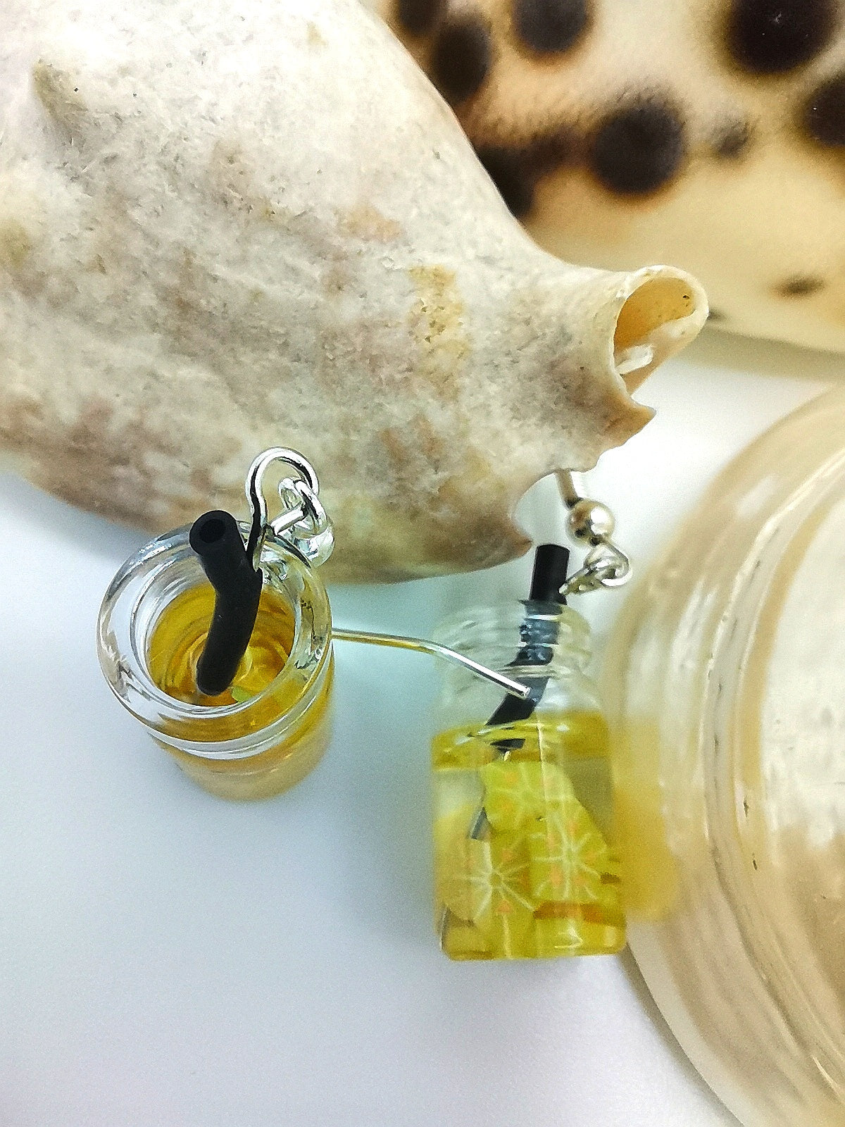 The Silver Sunbird Party Cocktail Earrings feature tiny glass jars resembling tropical drinks, one with a straw, next to a large seashell and shiny glass object. Filled with yellow liquid and summer resin fruits, they evoke a beach theme ideal for handmade earring inspiration.