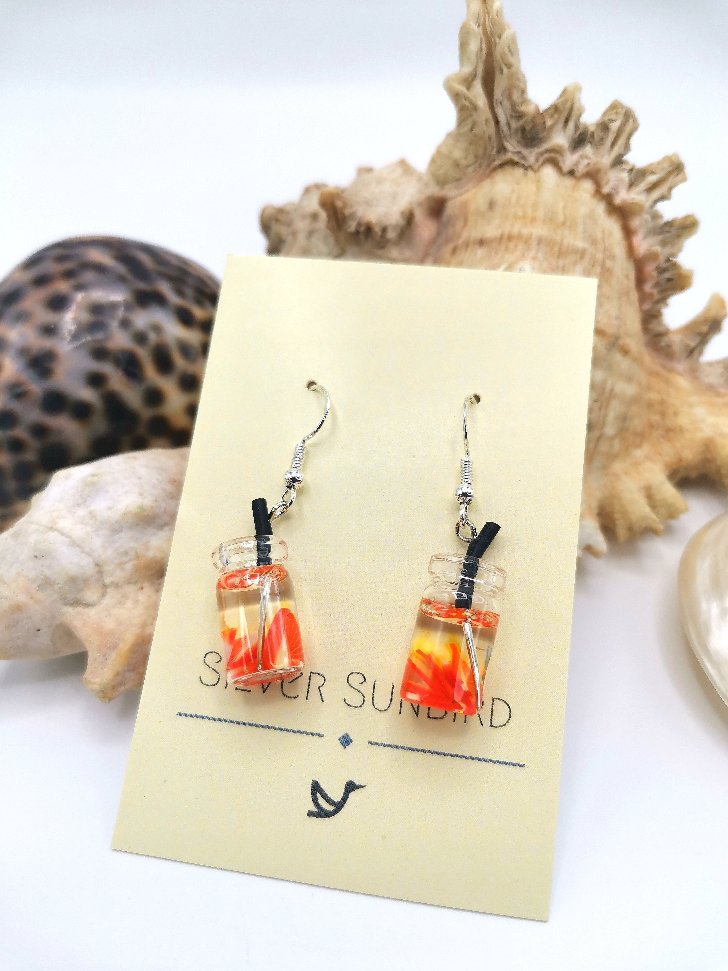 The Party Cocktail Earrings by Silver Sunbird are handmade, featuring small glass bottles with swirling orange patterns inside, sealed with corks. They hang from black cords against a card background with shells, embodying the essence of summer resin fruits.