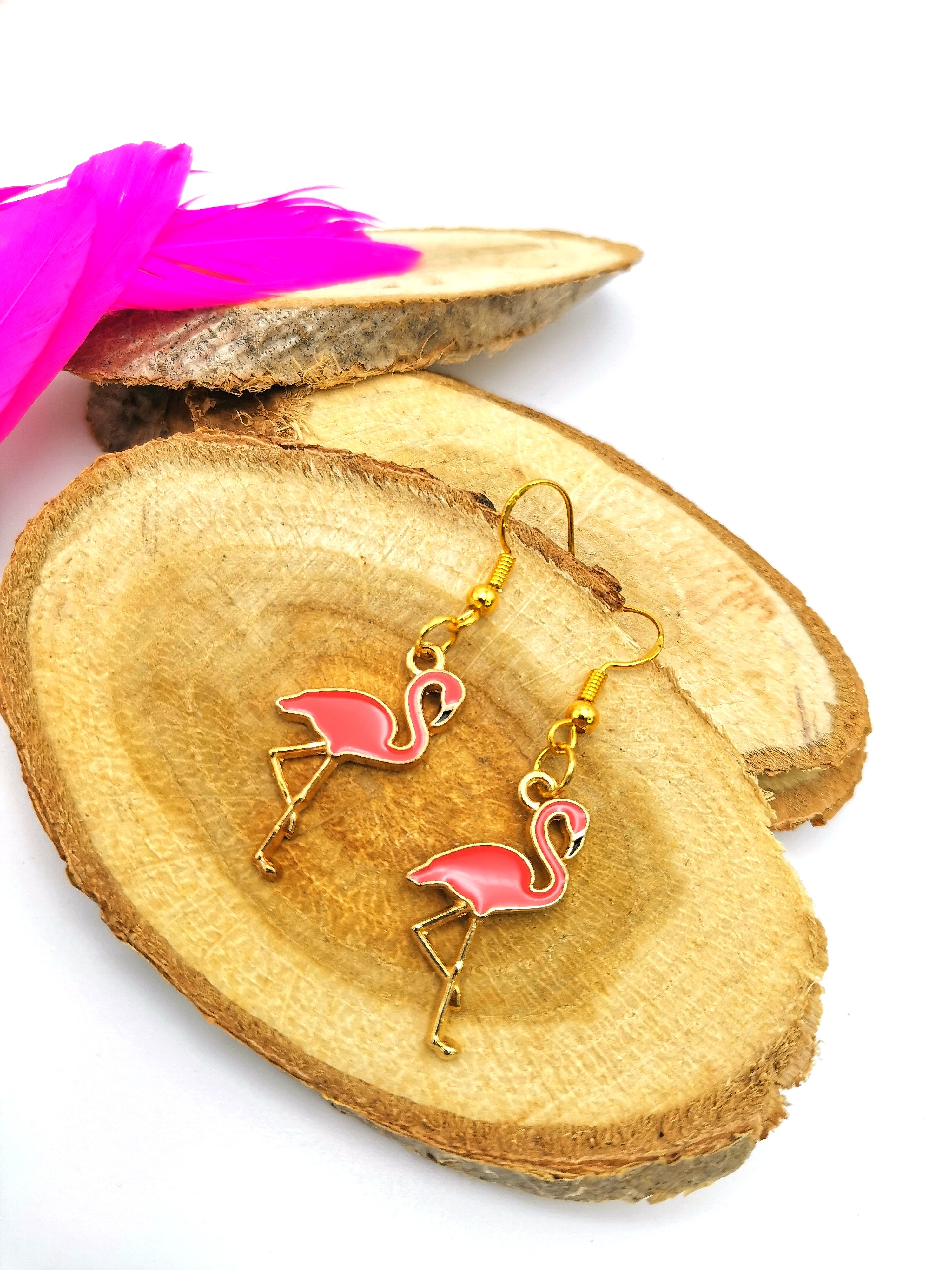 The Dancing Flamingo Earrings by Silver Sunbird, featuring a pink and gold color scheme, are displayed on a pair of round wooden slices. A bright pink feather adorns the top left corner. The white background accentuates the elegance of this handmade jewellery crafted from gold-plated sterling silver.