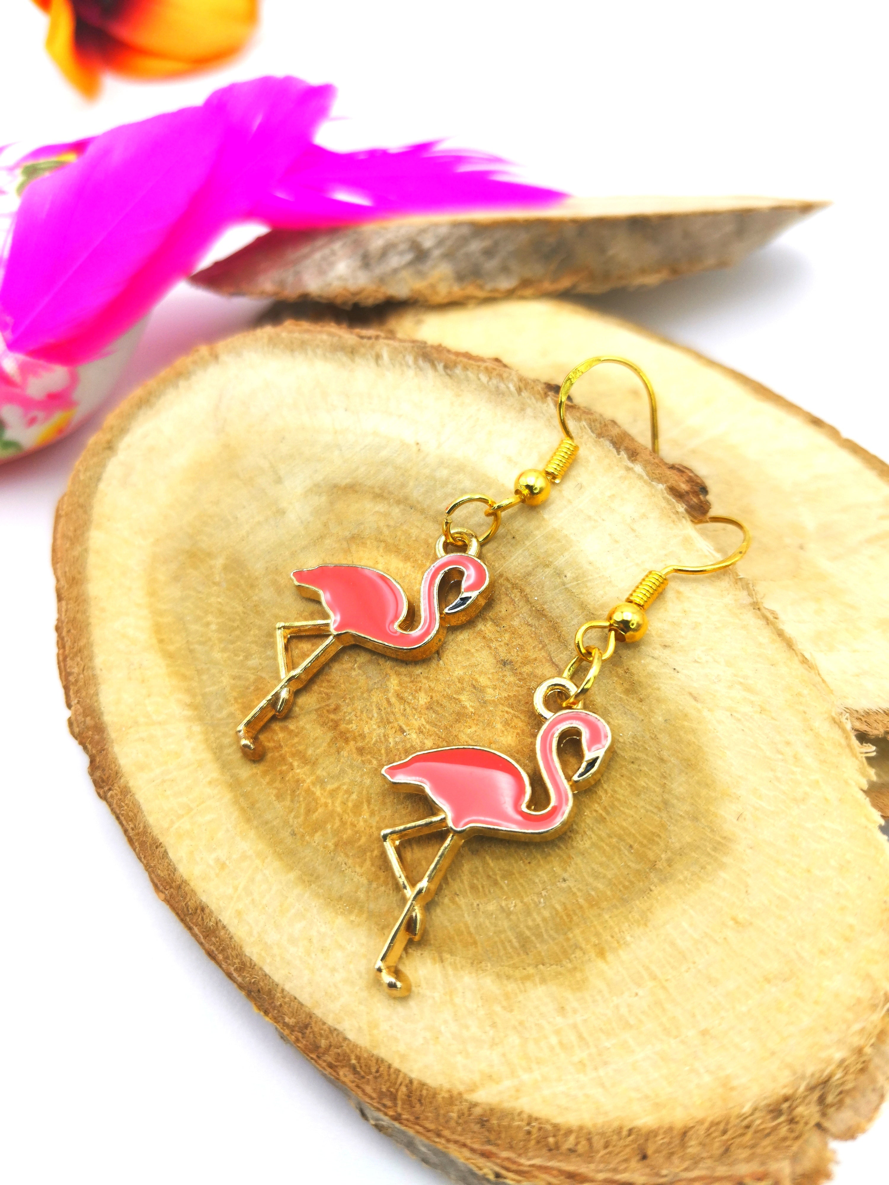 A pair of Dancing Flamingo Earrings by Silver Sunbird, crafted from gold-plated sterling silver and pink enamel, lie on a wooden slice. This handmade jewellery features flamingos with one leg lifted, detailed with gold outlines. The background also showcases vibrant pink and orange feathered accessories, adding a colorful touch.
