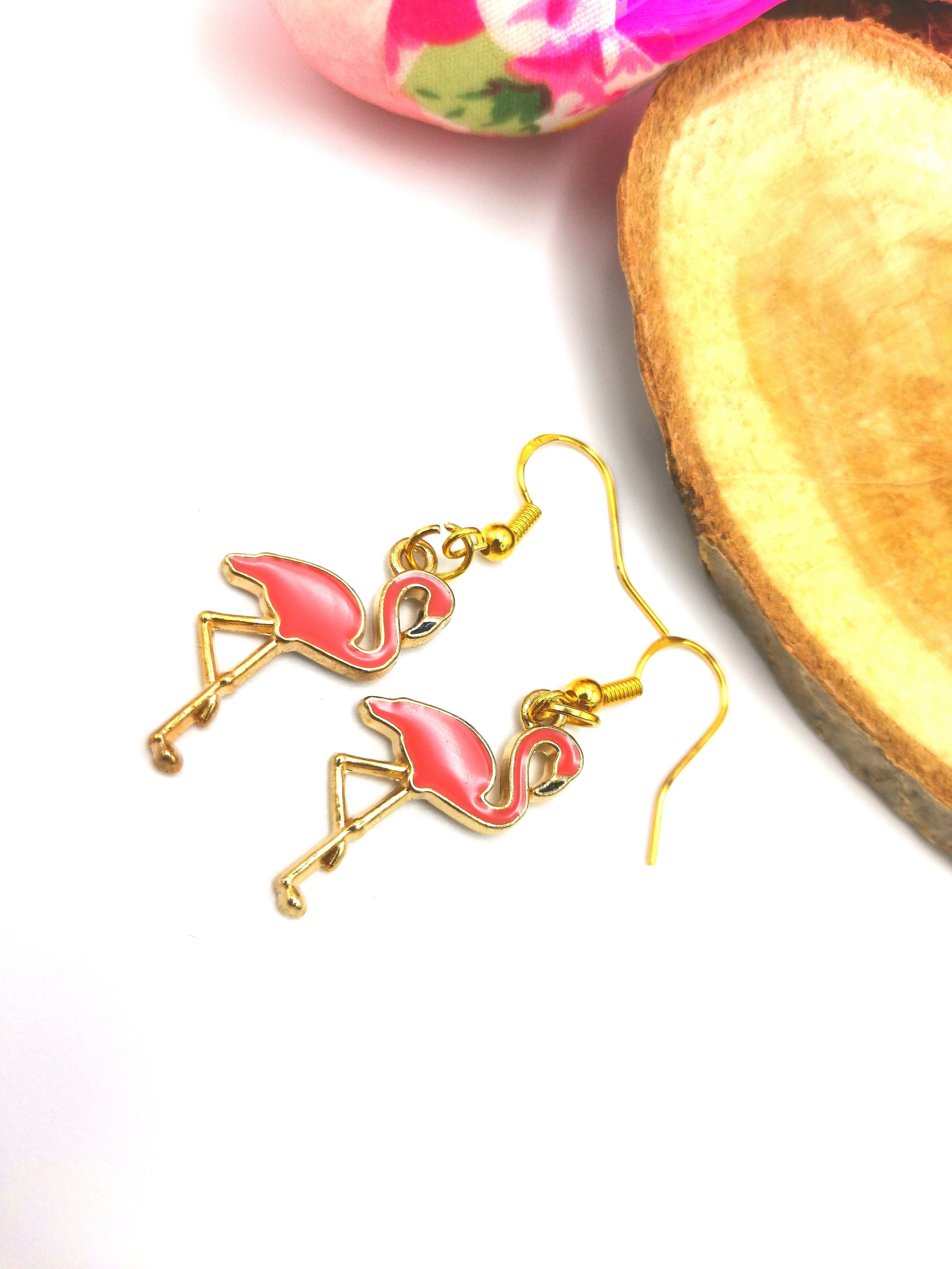 The "Dancing Flamingo Earrings" by Silver Sunbird, meticulously crafted from gold-plated sterling silver, are displayed on a white surface. Nearby, a vibrant pink floral fabric and a wooden item partially visible in the background add to the charm of this handmade jewellery scene.