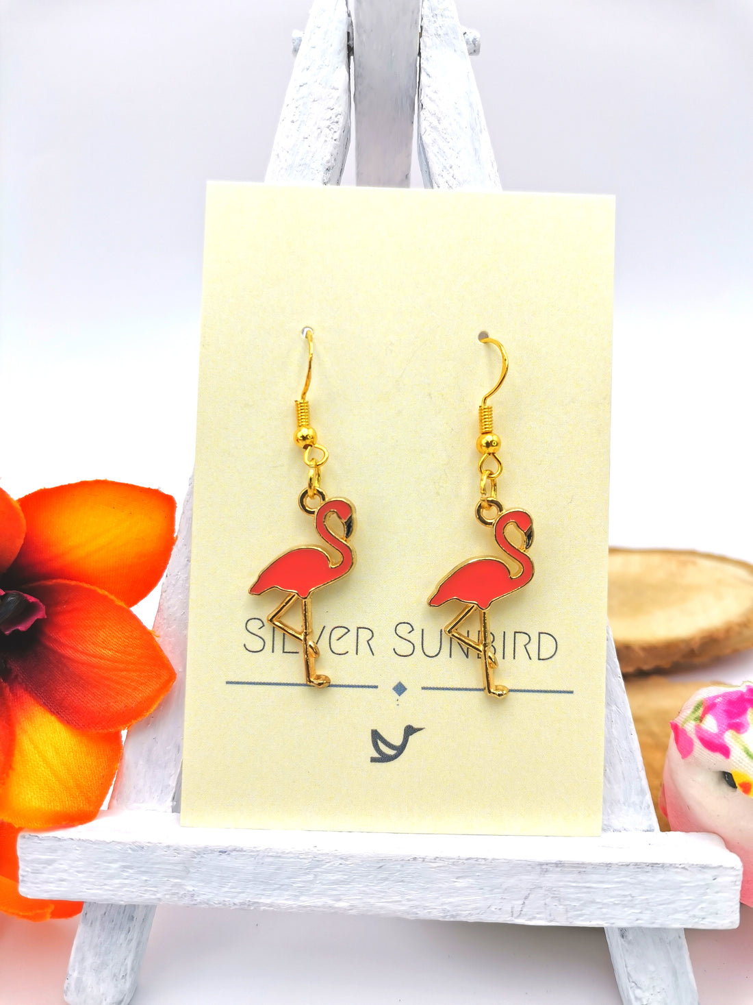 A pair of Silver Sunbird Dancing Flamingo Earrings with gold plated sterling silver hooks and pink flamingo pendants, elegantly displayed on a pale yellow-coloured card. The card is propped on a small easel, accompanied by a vibrant orange flower on the left and a small bowl with decorative items on the right, highlighting this exquisite piece of handmade jewellery.