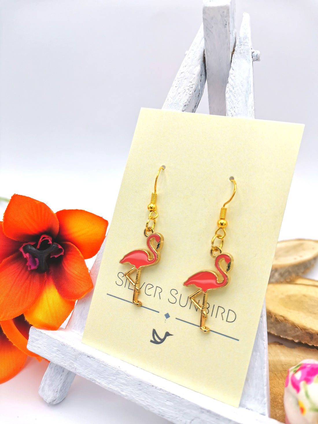 A display of handmade jewelry featuring the "Dancing Flamingo Earrings" with gold hooks and pink flamingo pendants hangs from a small white easel stand. Nearby, a vibrant orange flower accents the setup against a backdrop of wooden slices. The label proudly reads "Silver Sunbird".