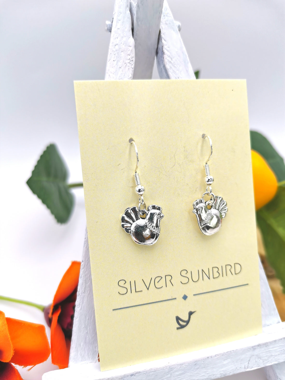 The Charismatic Chicken Earrings from Silver Sunbird, displayed on a card with the brand name, hang on a small easel surrounded by vibrant orange flowers and lush green leaves.
