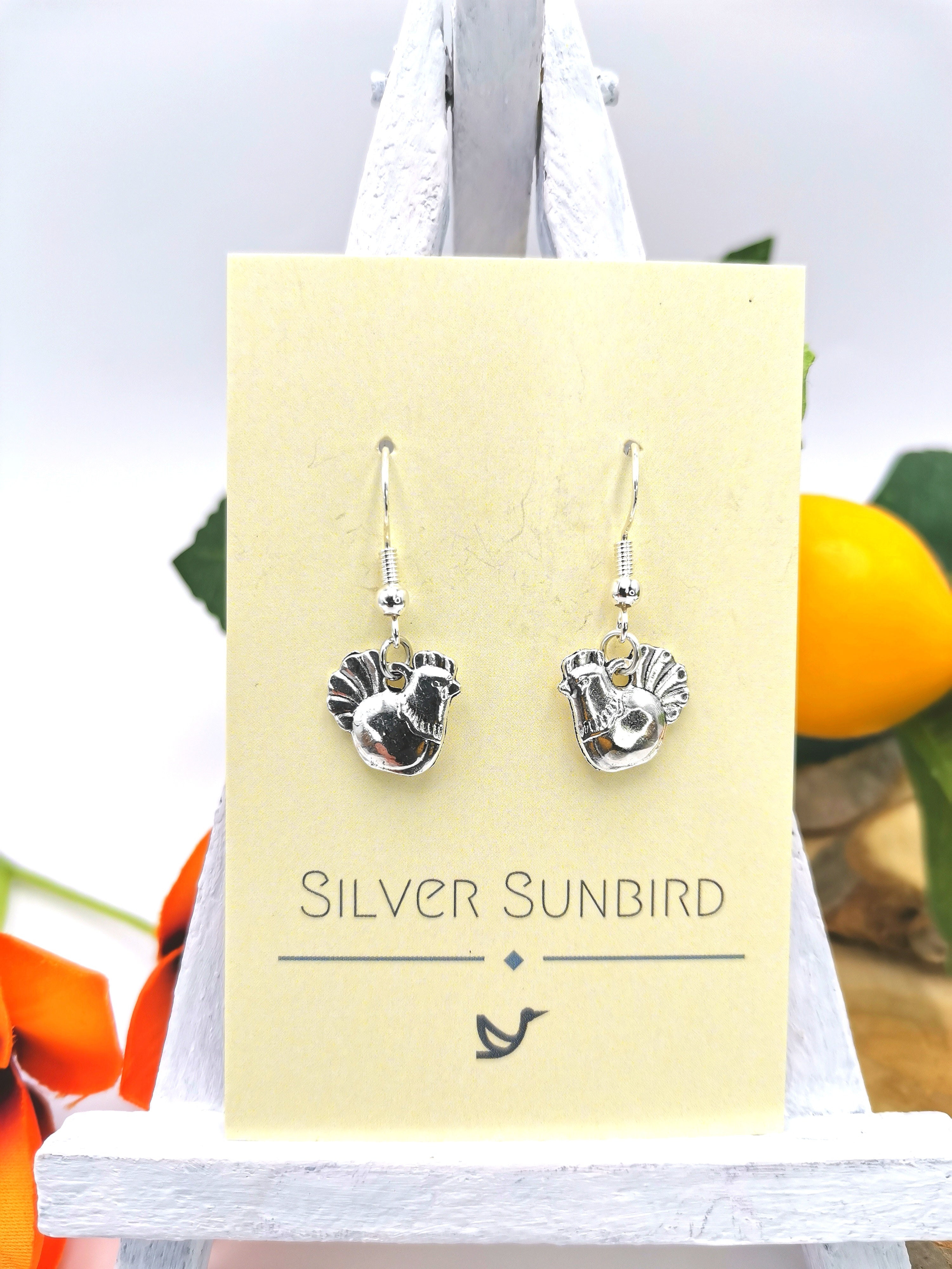 Sterling silver, rooster-shaped "Charismatic Chicken Earrings" by Silver Sunbird, displayed on a branded cardboard backing. These handmade earrings showcase intricate detailing against a blurred orange and green background.