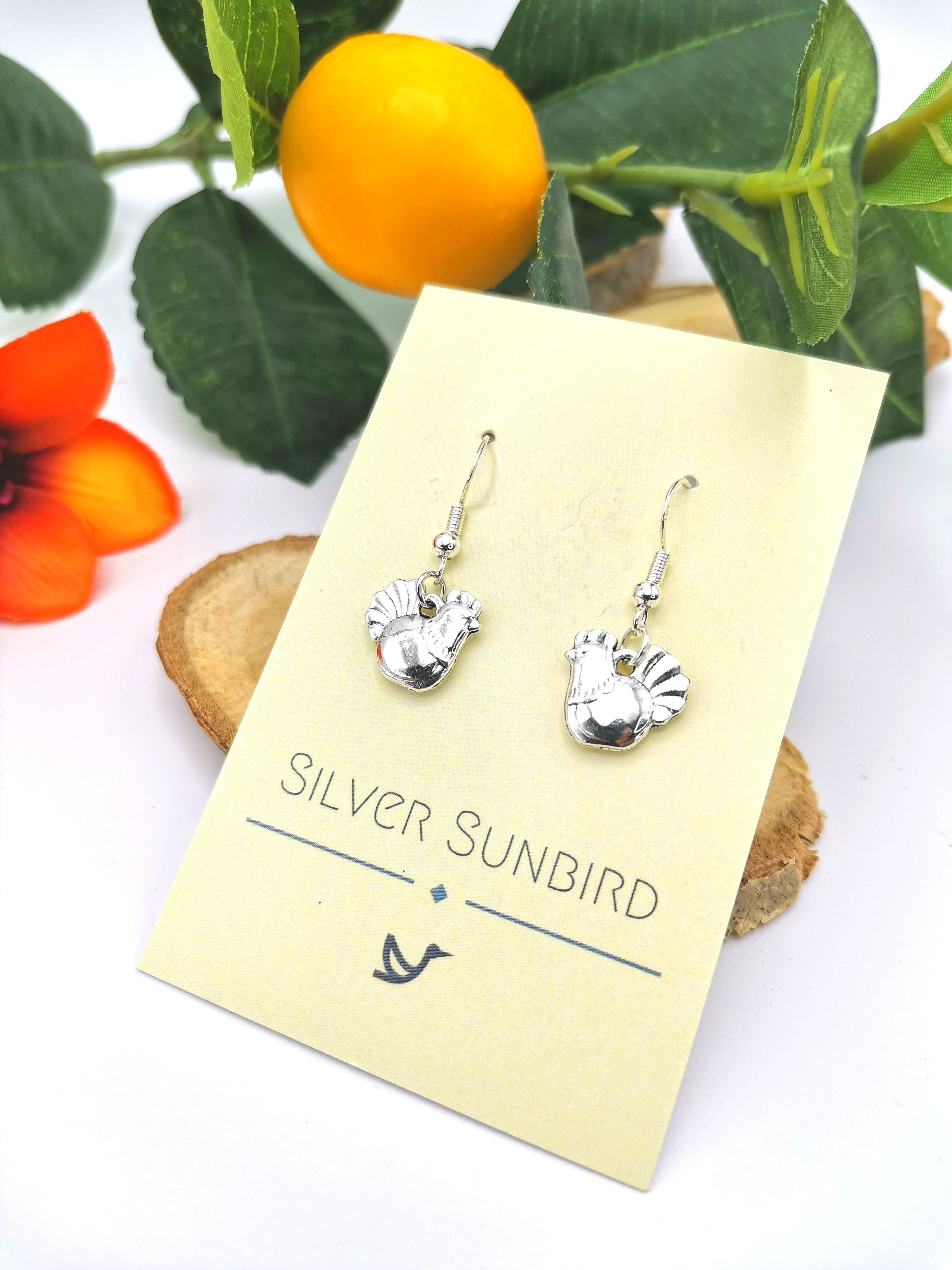 A pair of handmade Charismatic Chicken Earrings is displayed on a Silver Sunbird card, set against wooden slices, a vibrant orange flower, and green leaves with an orange fruit.
