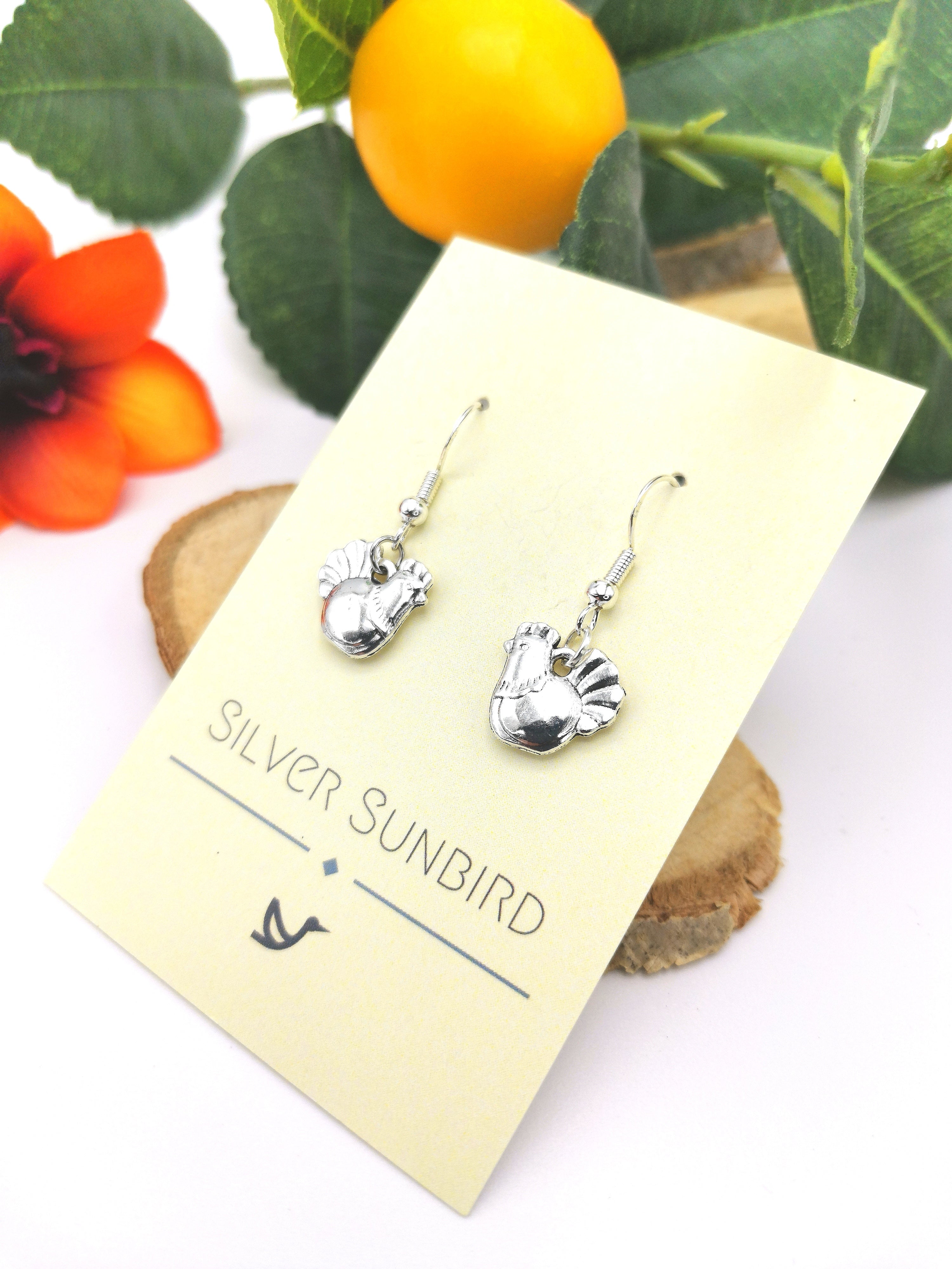Charismatic Chicken Earrings by Silver Sunbird are showcased on a card, set against bright orange flowers and green leaves. Crafted from sterling silver in a bird shape, they bring a natural vibrancy to the scene.
