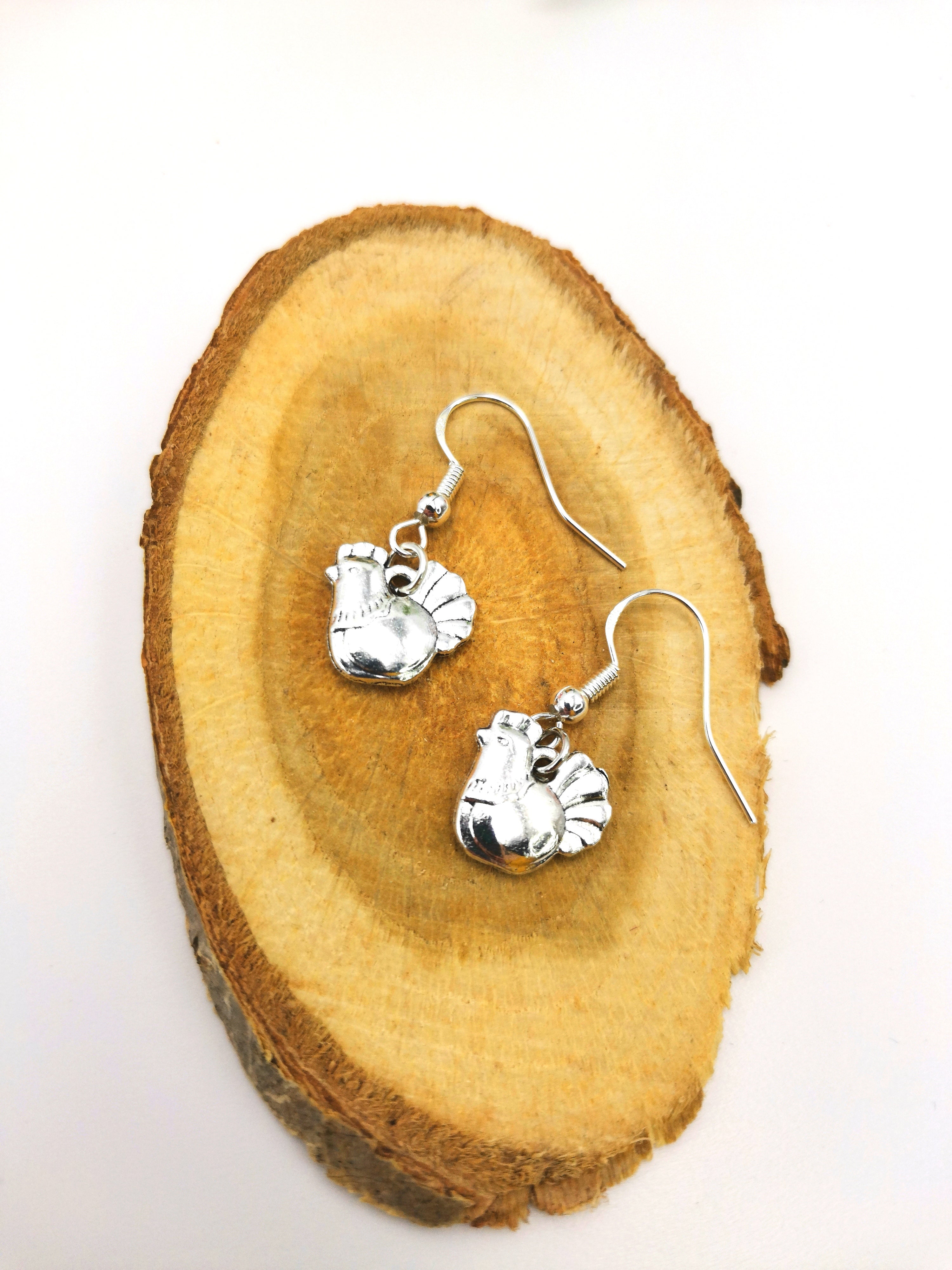 The Charismatic Chicken Earrings by Silver Sunbird, a handmade pair of sterling silver rooster-shaped earrings, is displayed on a round wooden slice with a light background.