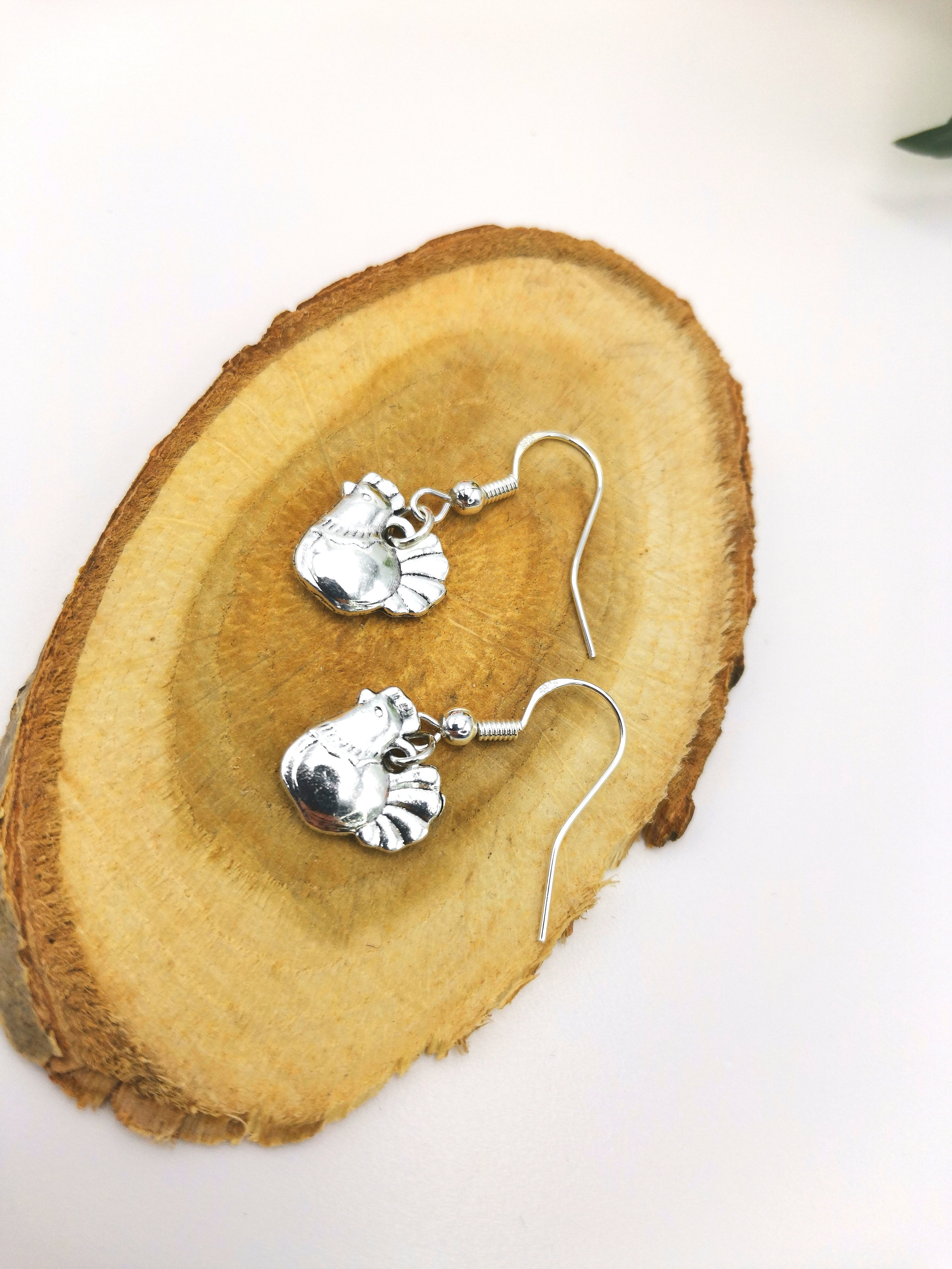 Charismatic Chicken Earrings by Silver Sunbird are handmade sterling silver earrings featuring intricate chicken designs. Hanging from hooks, they rest on a wooden slice backdrop, adding a rustic touch.