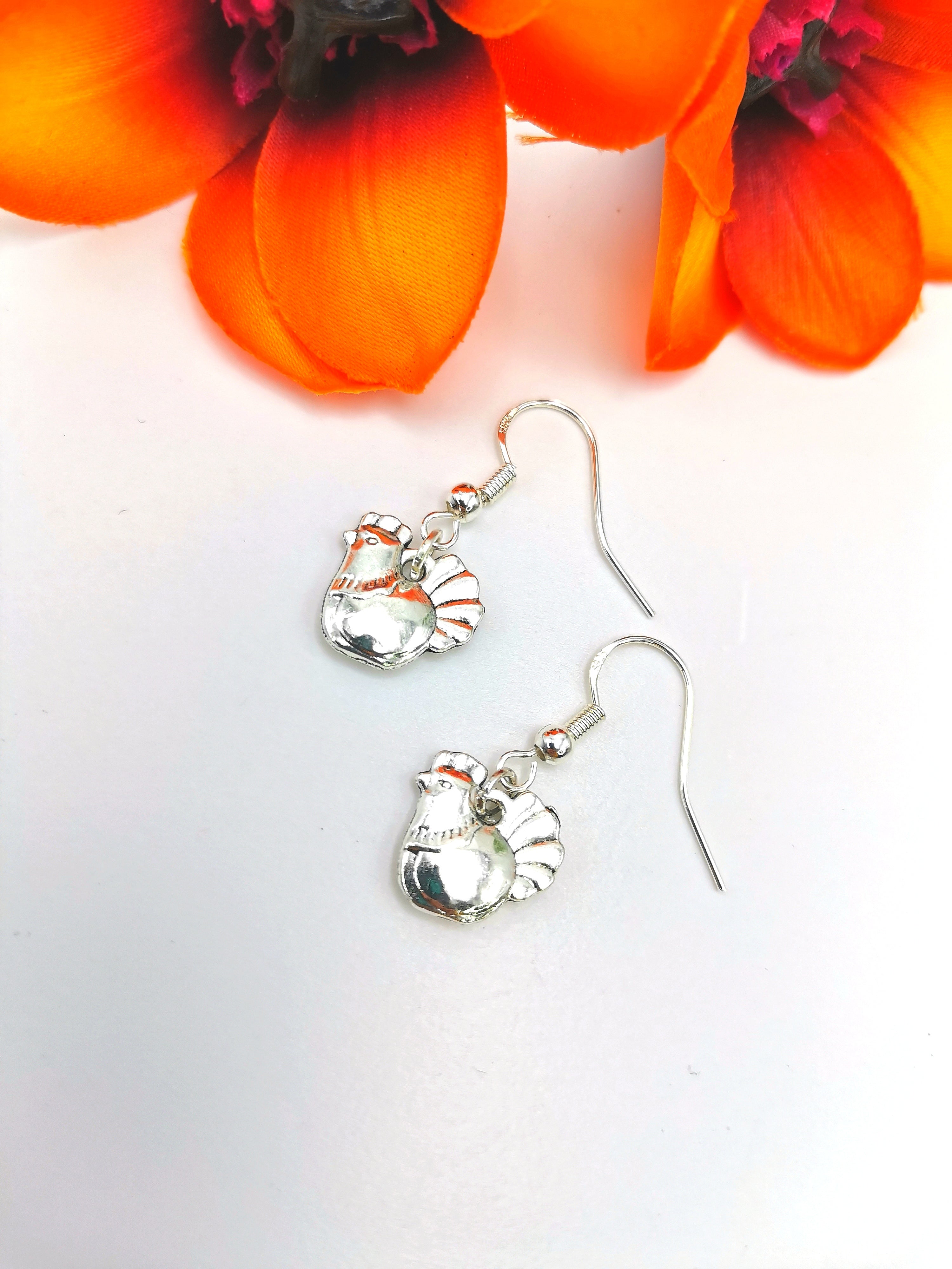 A pair of handmade Charismatic Chicken Earrings by Silver Sunbird rests on a white surface, flanked above by vibrant orange flowers, adding warm contrast to the shiny sterling silver.
