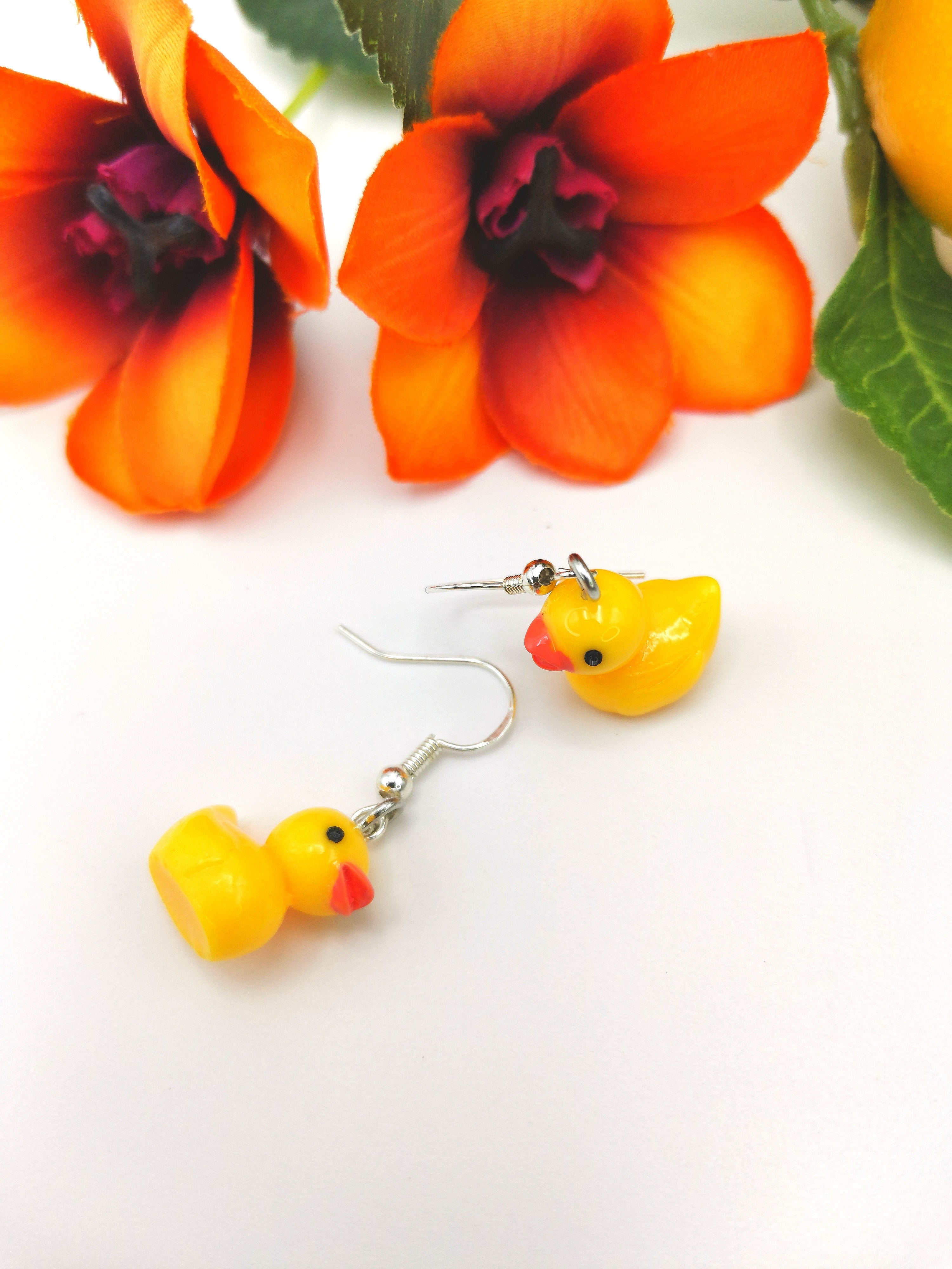 The Silver Sunbird Young Hearted Yellow Ducks Earrings are showcased on a white surface, paired with two vibrant orange flowers in the background for a playful touch.