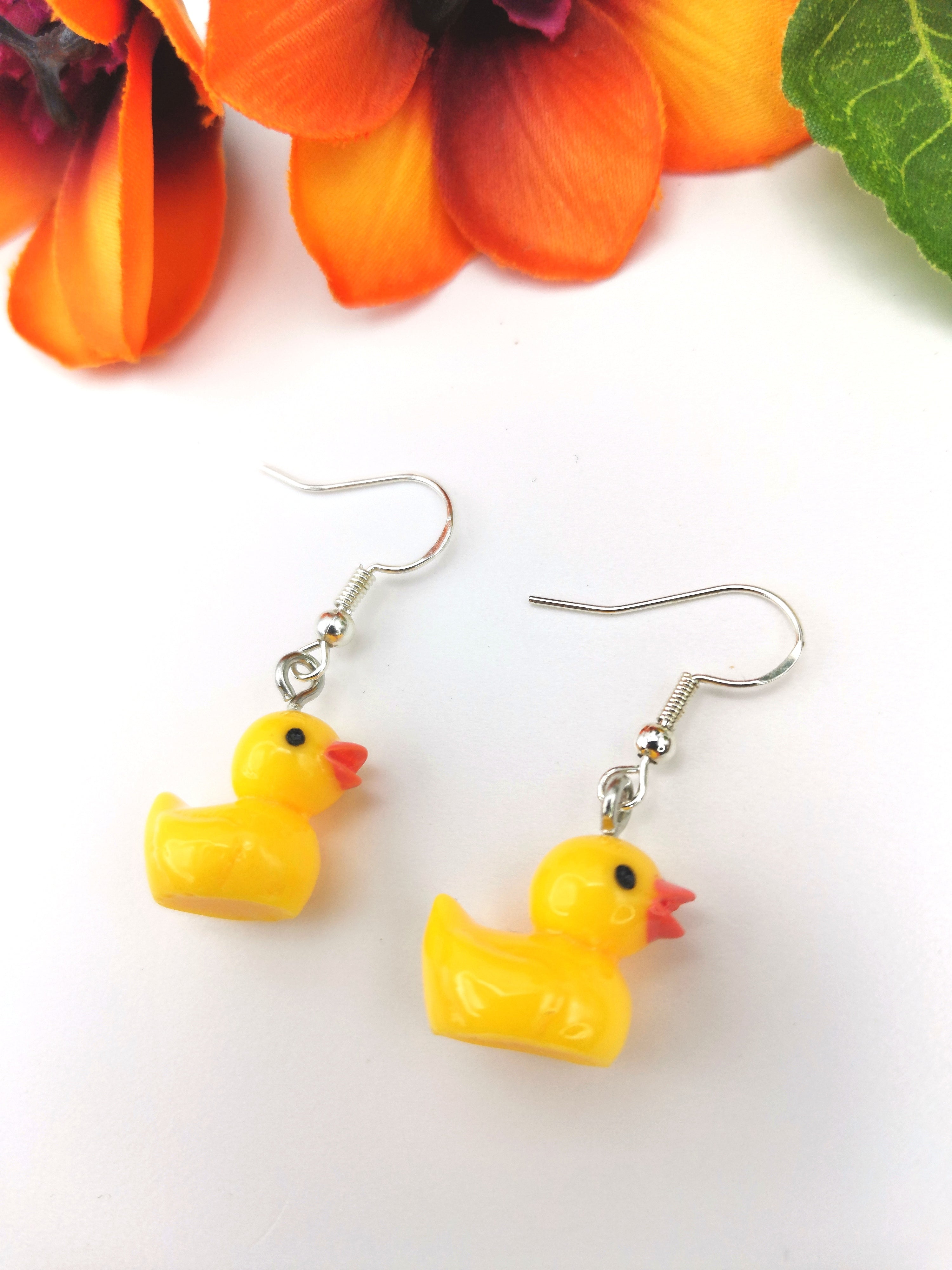 The Silver Sunbird Young Hearted Yellow Ducks Earrings dangle against a white background with orange flowers and a green leaf—fun, quirky jewelry to add playful style to your collection.