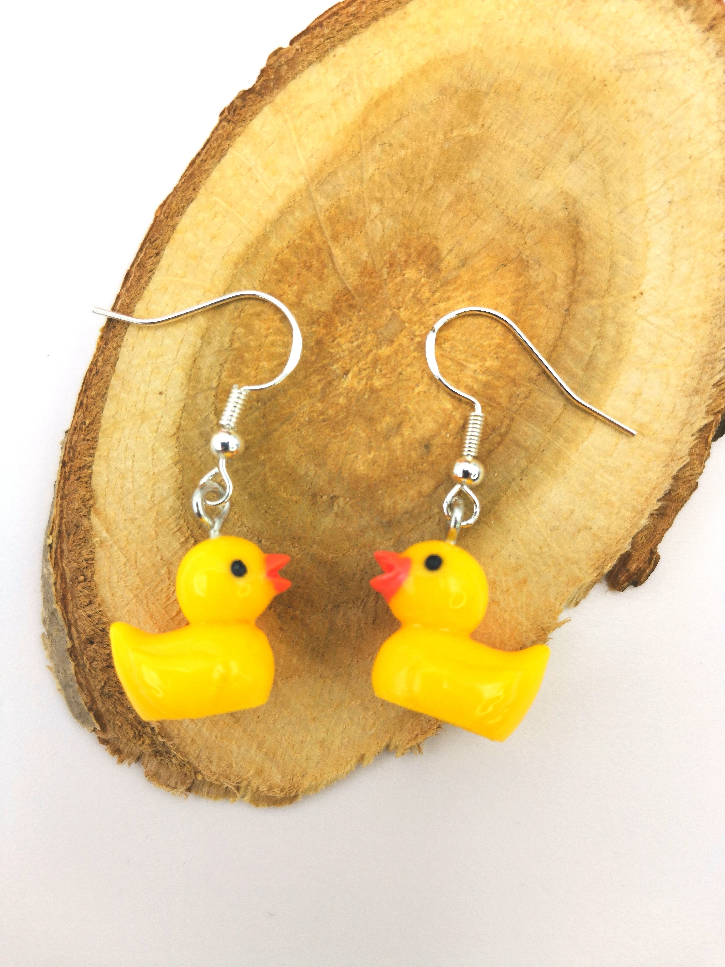 The Silver Sunbird Young Hearted Yellow Ducks Earrings showcase quirky, small yellow rubber duck charms, displayed on a light wooden surface with a white background.