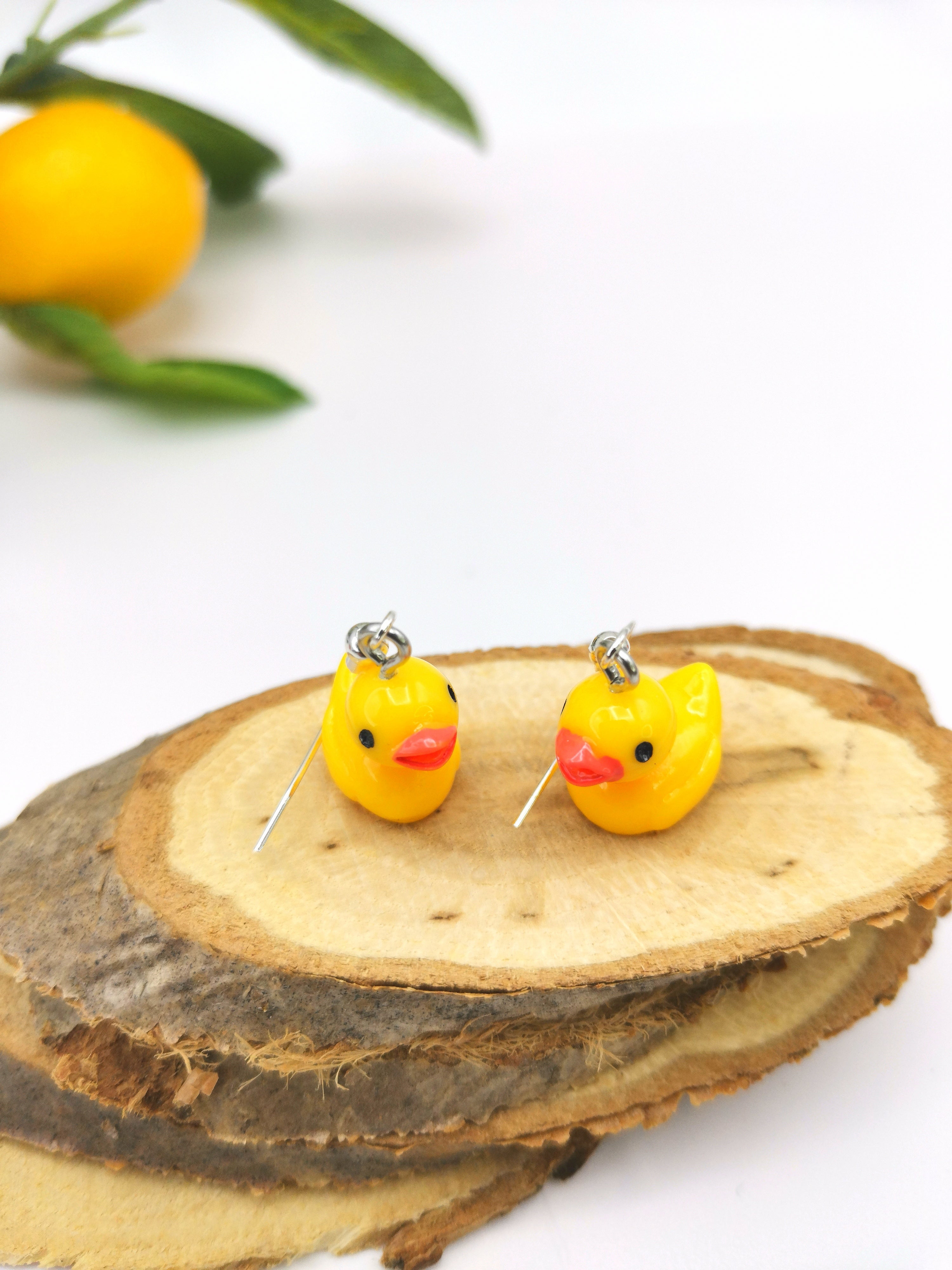 The Silver Sunbird Young Hearted Yellow Ducks Earrings, perfect for quirky jewellery lovers, are shown on wooden slices with a blurred orange and green leaves in the background, adding a playful touch to the bright, minimal setting.