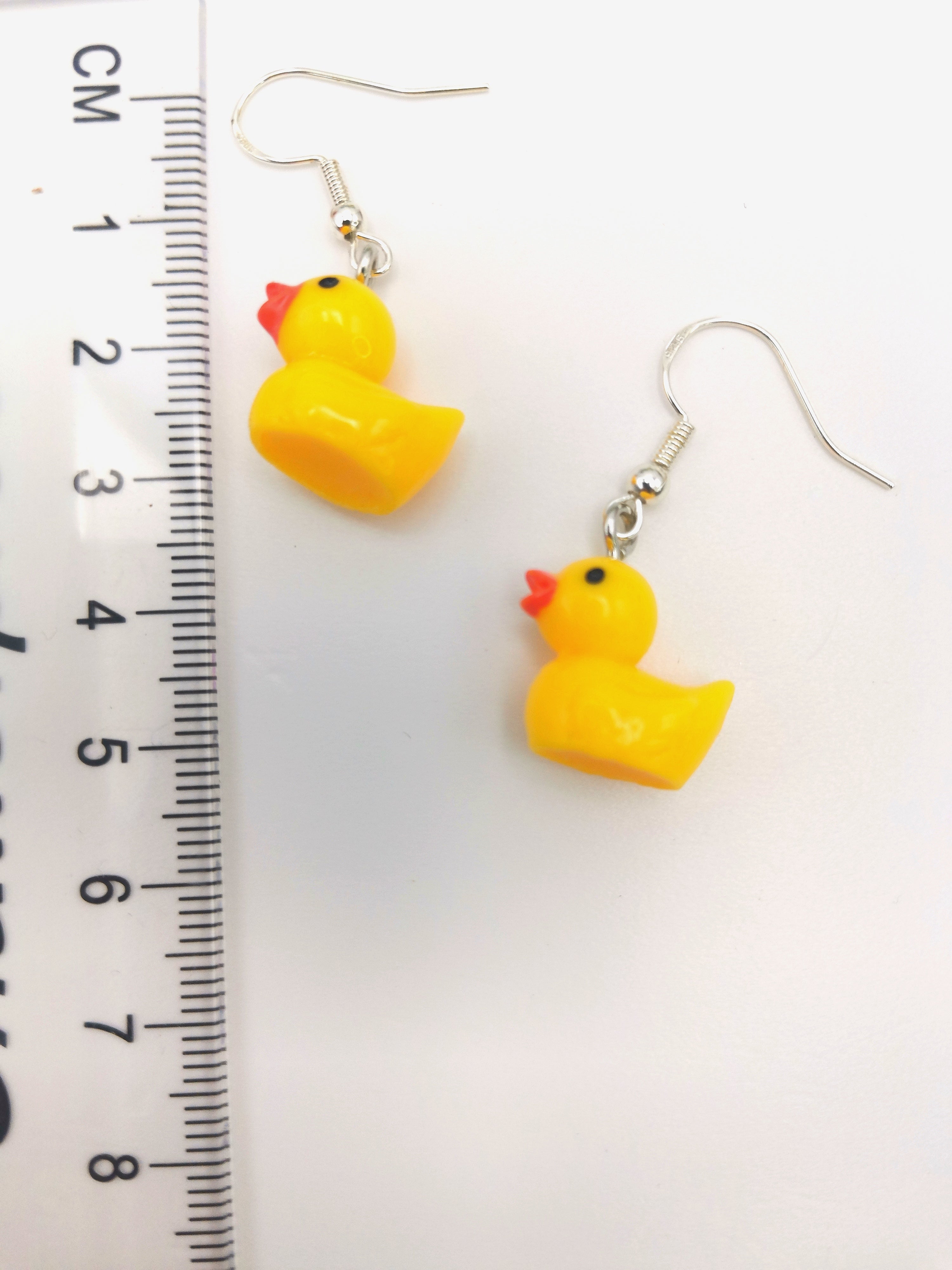 The Silver Sunbird Young Hearted Yellow Ducks Earrings feature a playful yellow duck design with orange beaks and silver hooks, shown beside a centimeter ruler on a white background.