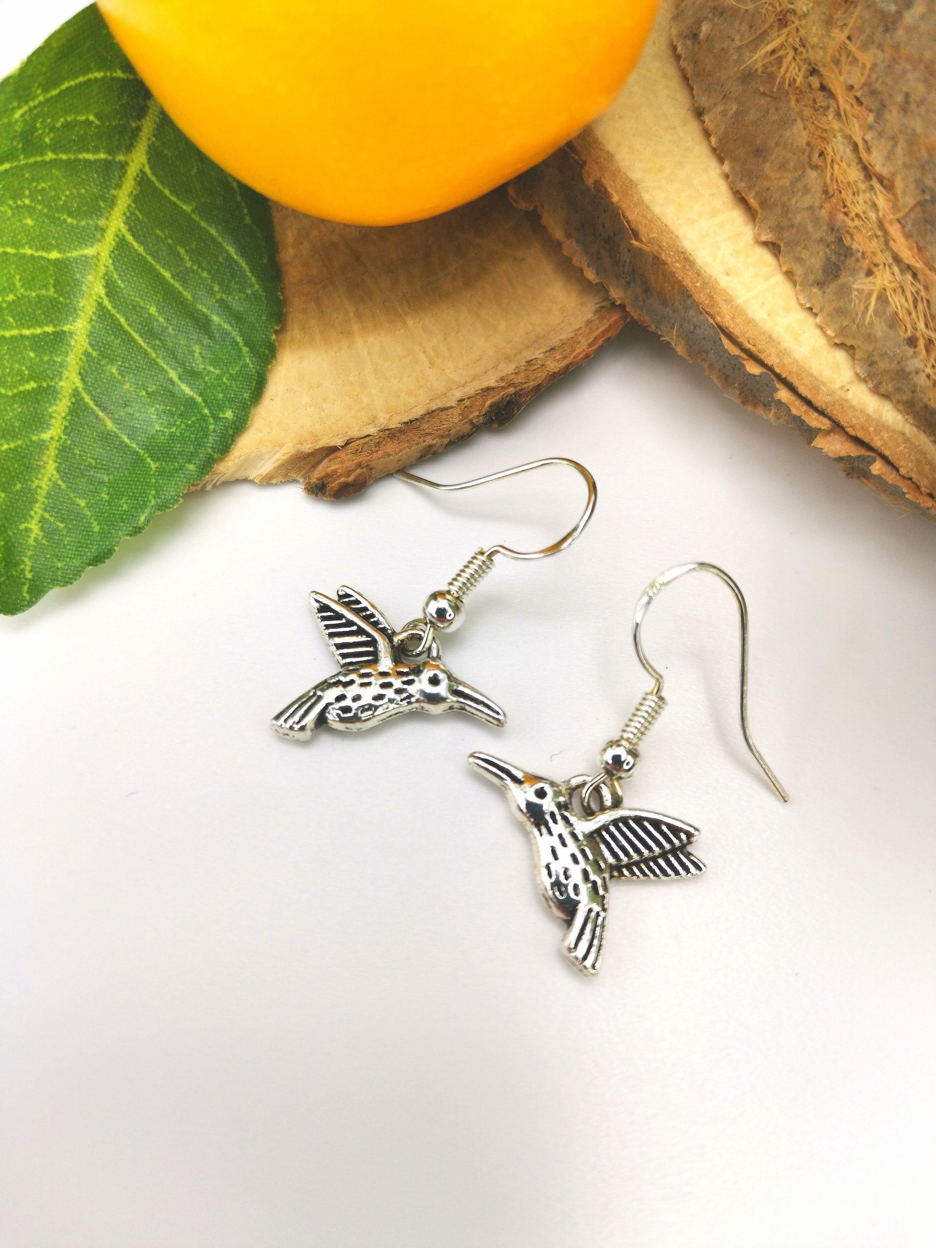 A pair of Heartfelt Hummingbird Earrings by Silver Sunbird is displayed on a white surface. The handmade jewellery features detailed hummingbird charms and is placed near a green leaf, a yellow-orange fruit slice, and a piece of bark.
