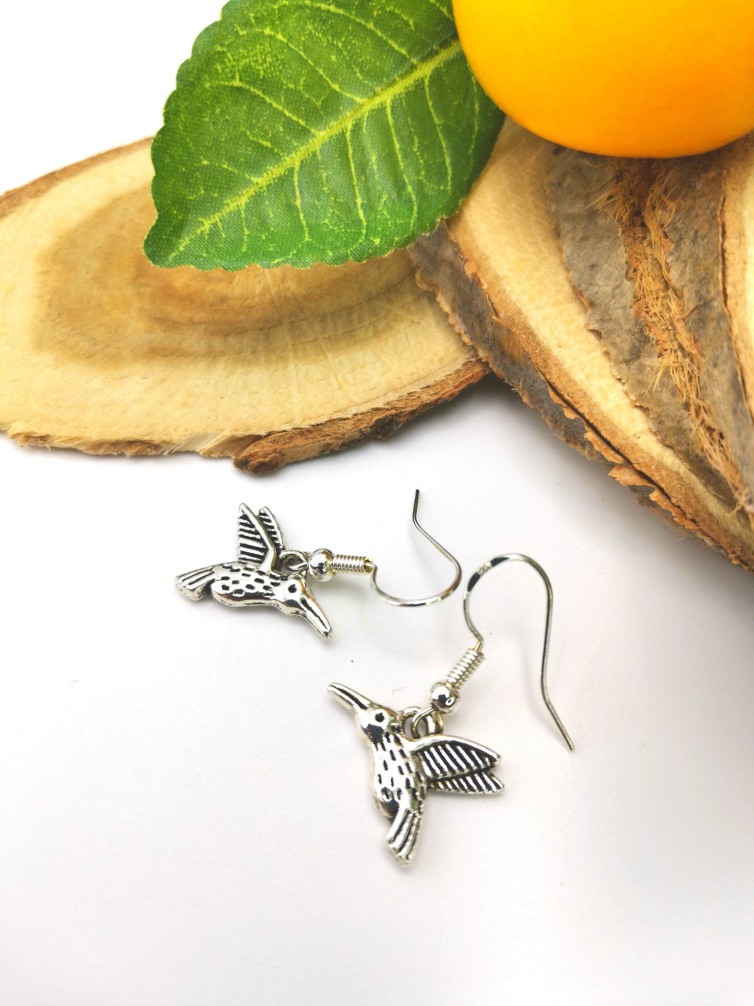 A pair of Heartfelt Hummingbird Earrings by Silver Sunbird with fishhook ear wires is placed on a piece of cut wood, accompanied by a green leaf and a yellow fruit. The delicate bird design features intricate details on the wings and body, showcasing the craftsmanship of handmade jewellery.