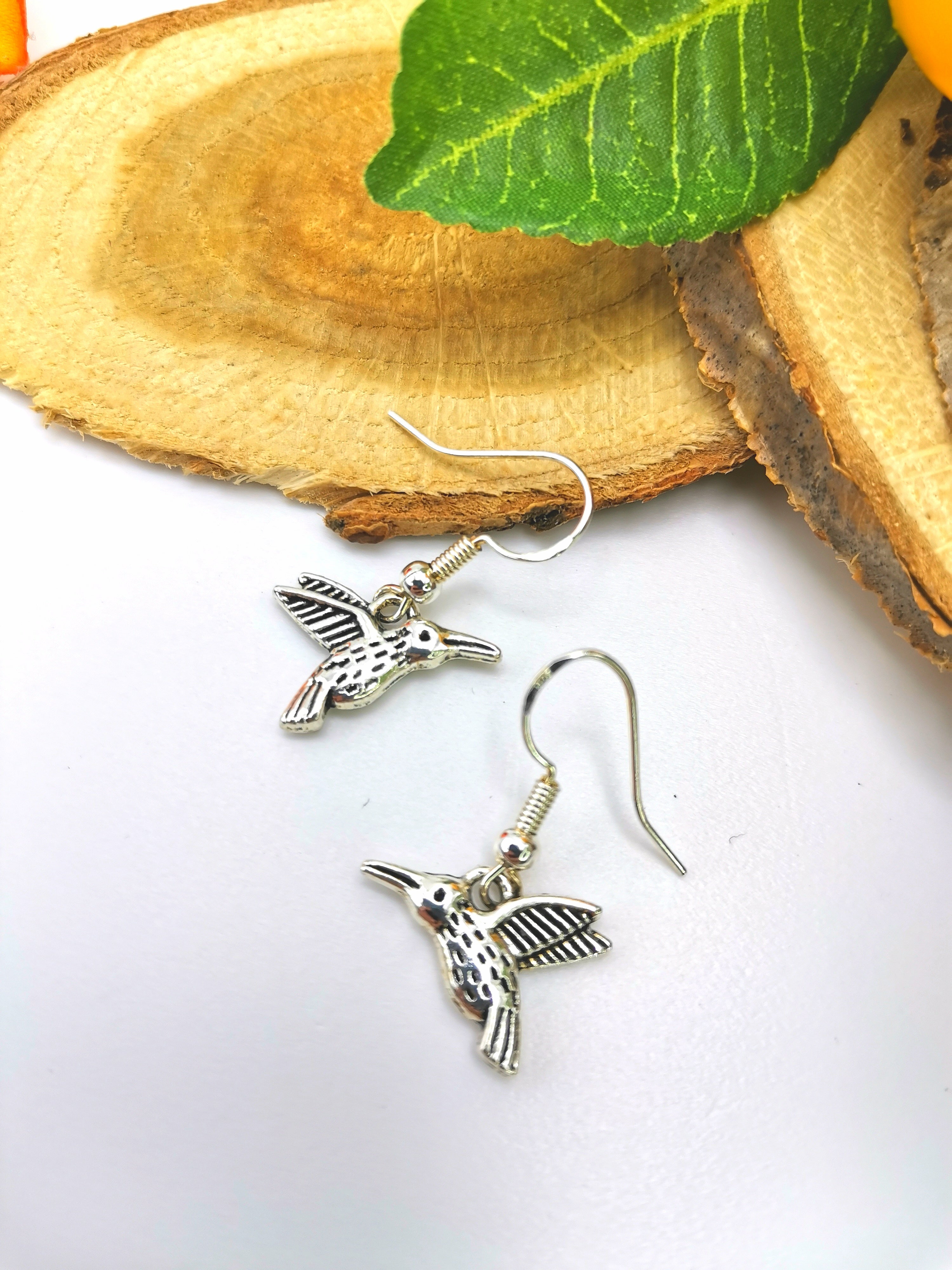 The Heartfelt Hummingbird Earrings by Silver Sunbird are meticulously handcrafted sterling silver pieces, featuring intricate detailing. Displayed on a wooden slice with a green leaf in the backdrop, these earrings have fish hook backings and showcase the wood&