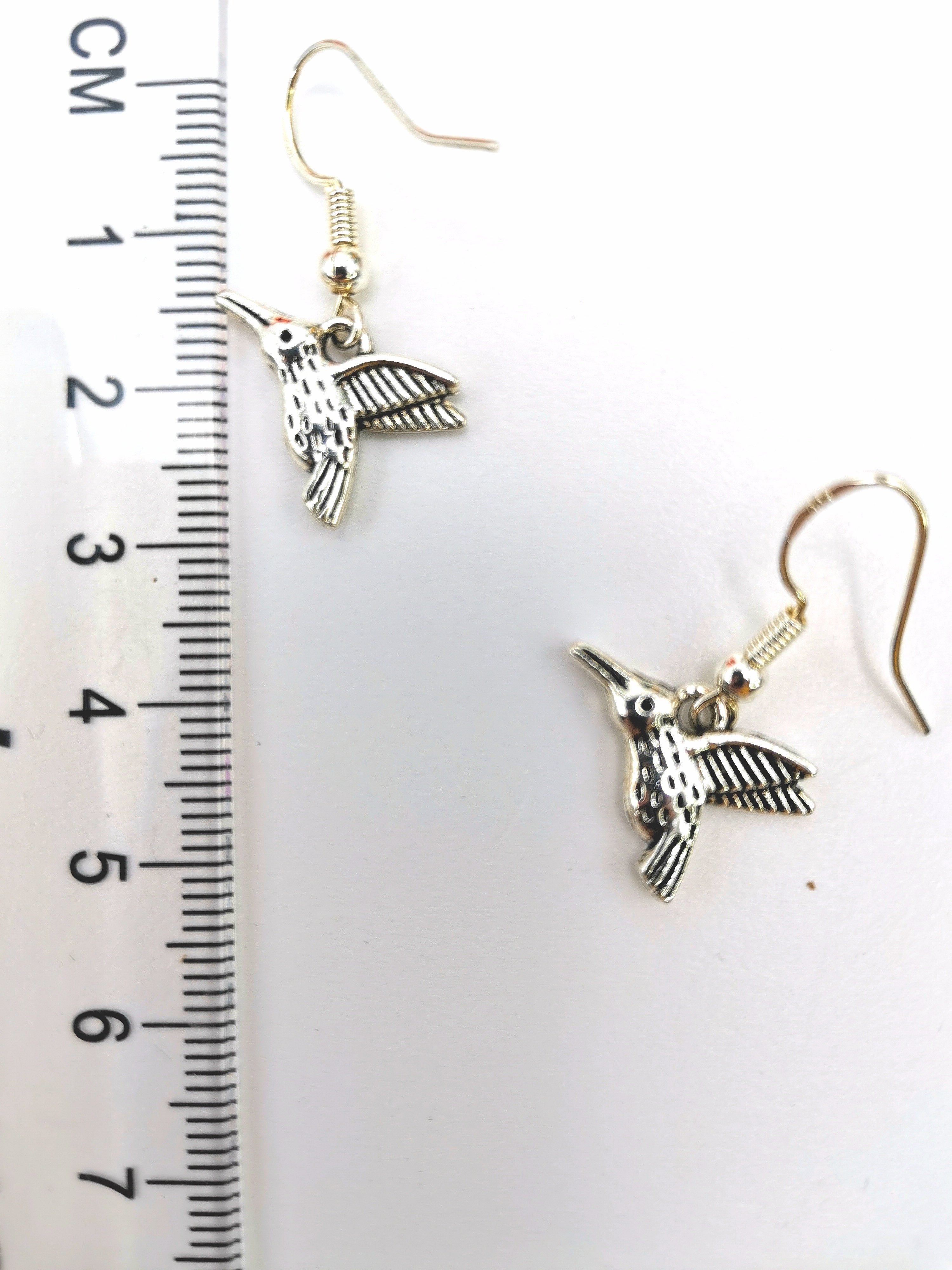 The Heartfelt Hummingbird Earrings by Silver Sunbird, meticulously handmade from sterling silver and designed in the shape of hummingbirds, are displayed next to a ruler on a white surface. The ruler indicates that each earring is about 2.5 cm long, emphasizing their delicate size and intricate details.