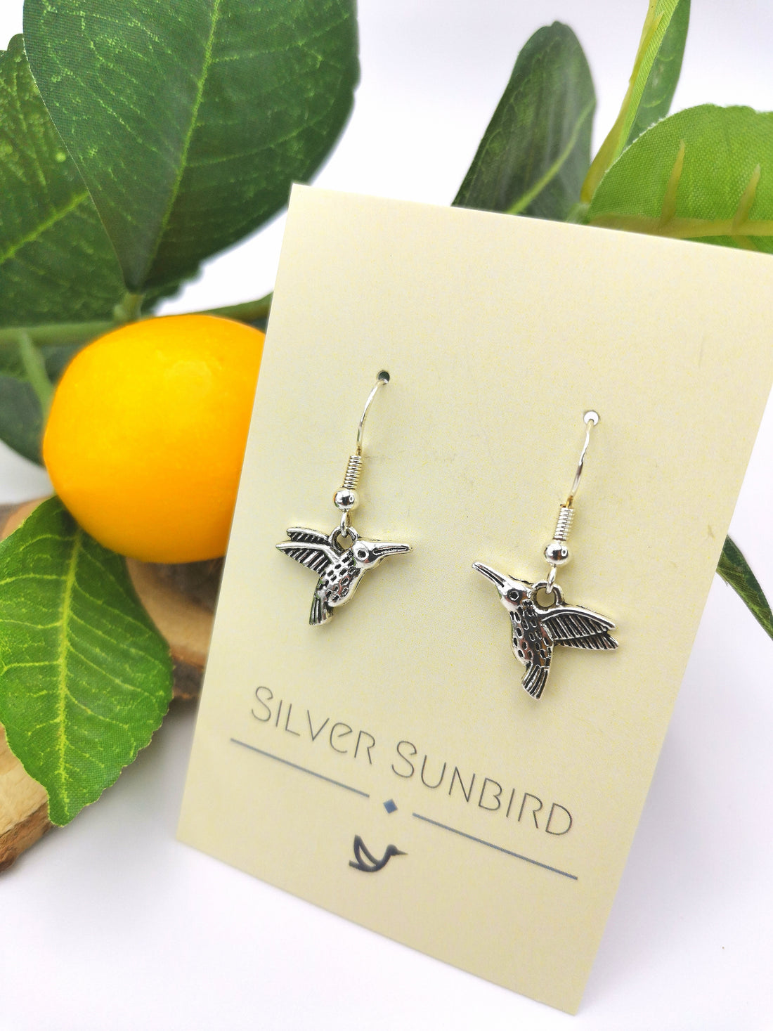 A pair of Heartfelt Hummingbird Earrings, meticulously crafted from sterling silver, displayed against a cream-colored card with "Silver Sunbird" printed on it. The card rests among green leaves and a small, yellow-orange fruit in the background.