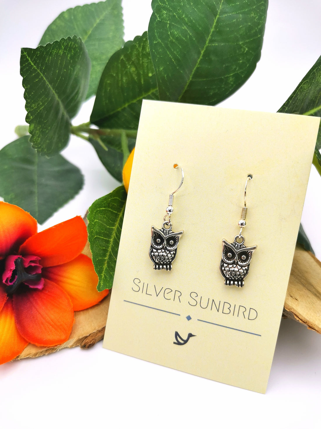 Enlightened Owl Earrings by Silver Sunbird on a wooden background with green leaves and an orange flower.