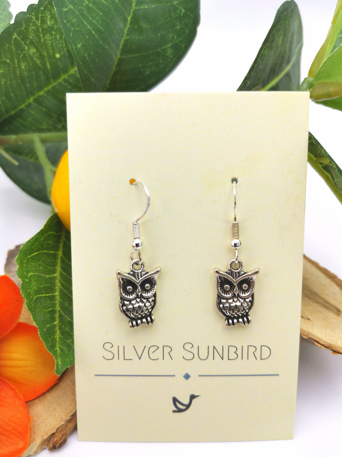 Enlightened Owl Earrings from Silver Sunbird gleam elegantly on a card set against lush green leaves and vibrant orange flowers.