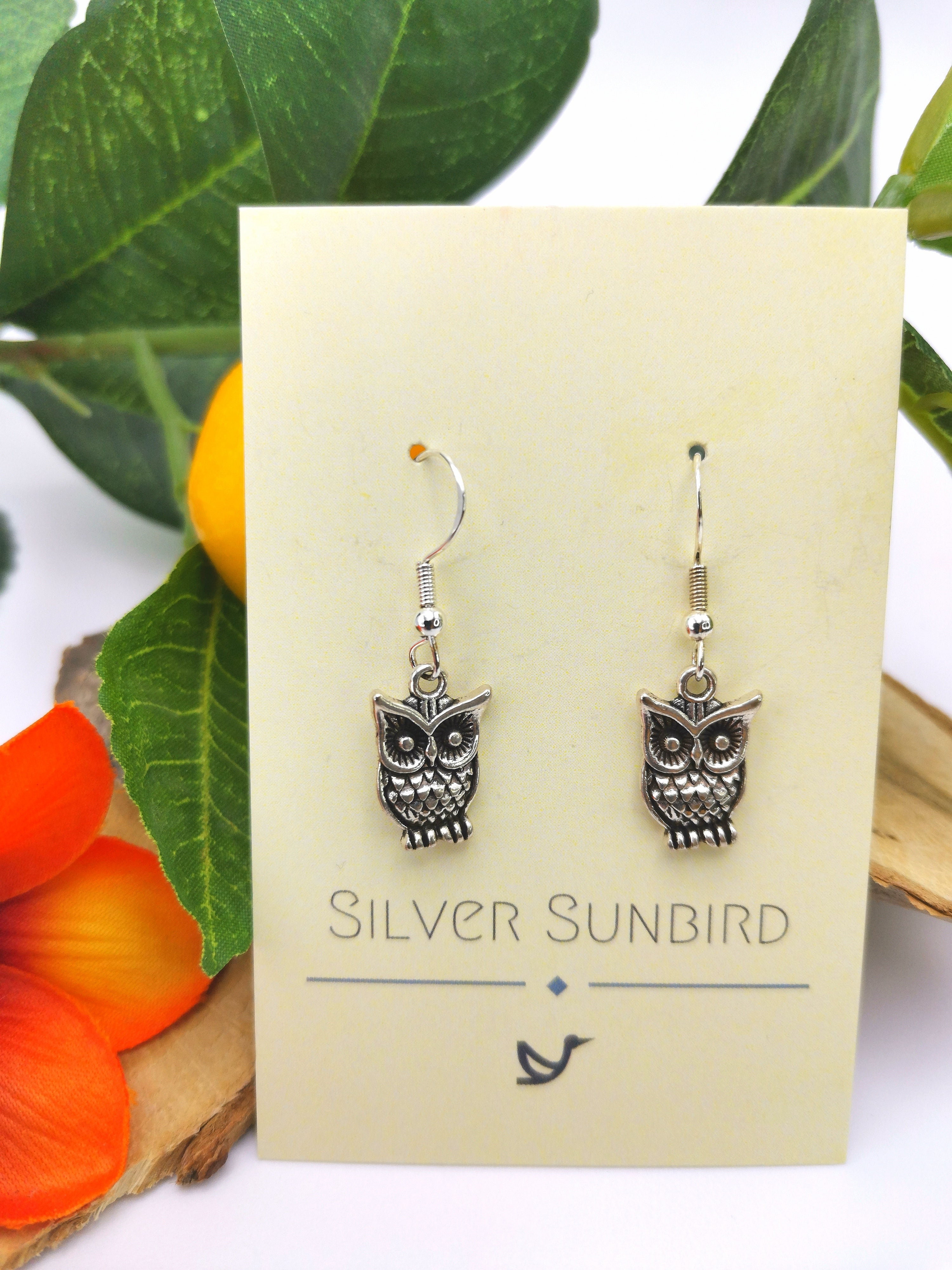 These sterling silver earrings, featuring enchanting owl charms, are displayed on a "Silver Sunbird" card. The backdrop of green leaves and vibrant orange-red flowers adds to the allure of these Enlightened Owl Earrings.