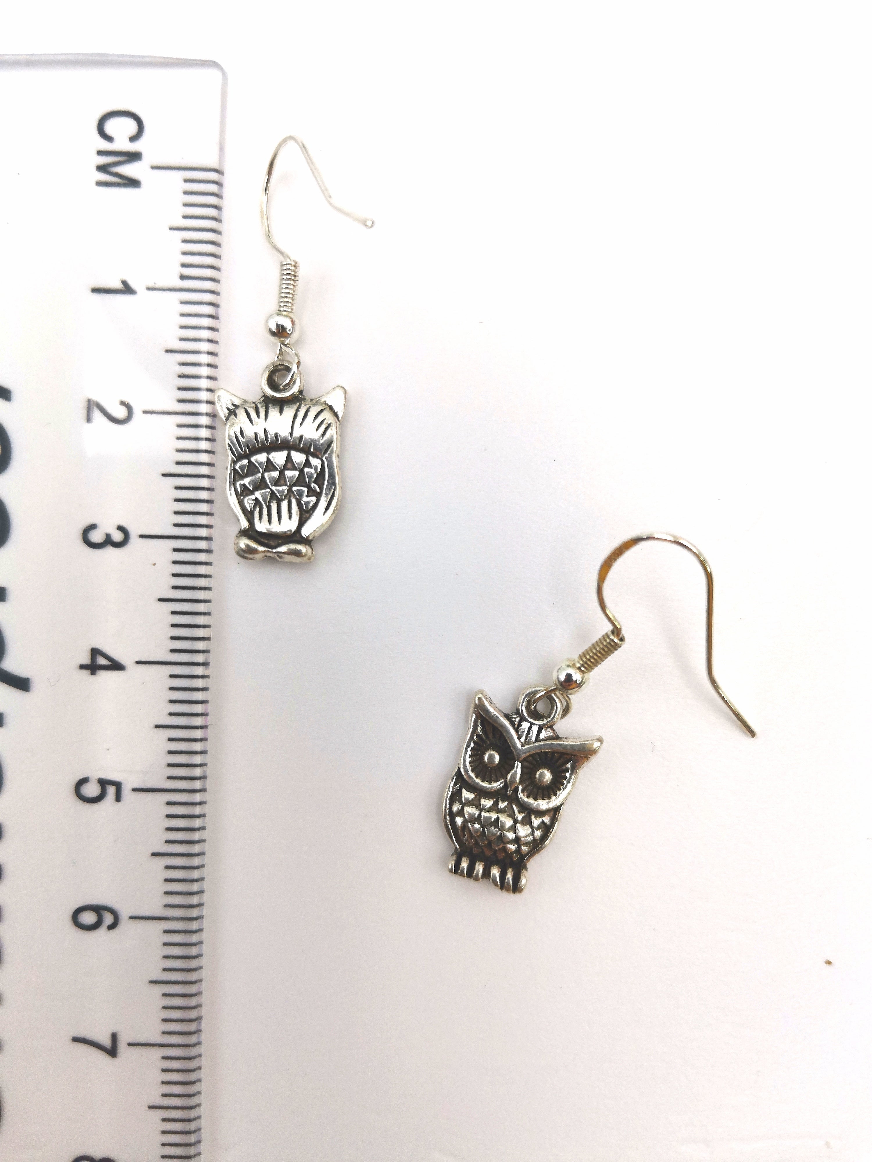 The Enlightened Owl Earrings by Silver Sunbird feature intricate owl designs and are crafted in sterling silver. Placed next to a ruler for scale, these charms showcase exquisite craftsmanship.